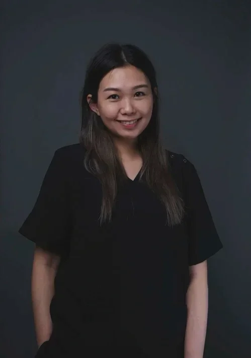 A young woman with long dark hair, smiling, wearing a black short-sleeved top, standing against a plain dark background.