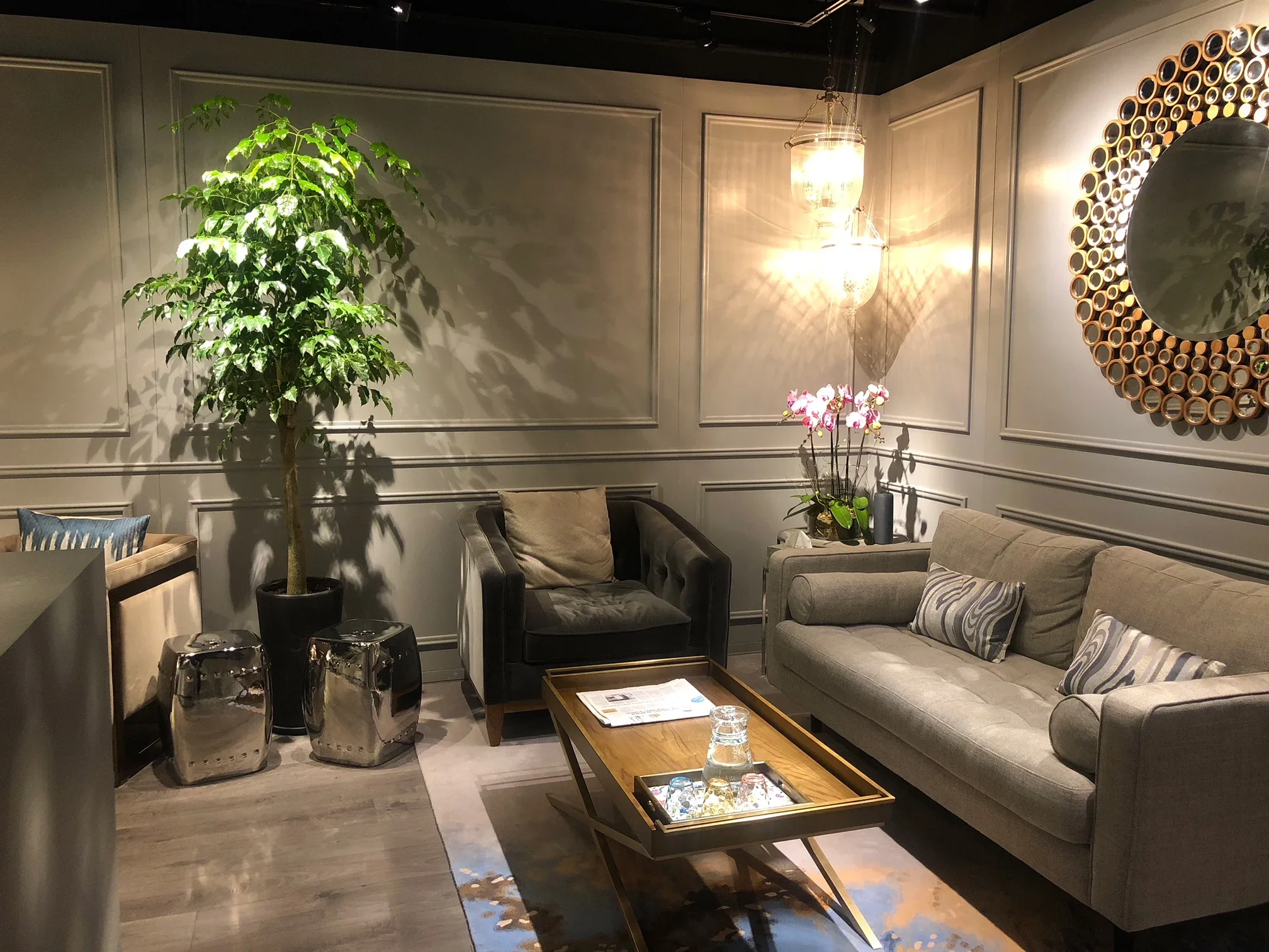 A cozy living room with a tall potted plant, beige sofas with striped pillows, a wooden coffee table with glasses and a tray, a wall mirror with circular frame, a modern chandelier, and metallic stools.