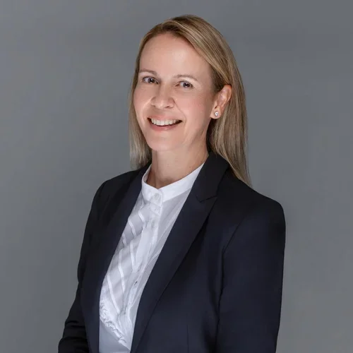 A woman with blonde hair wearing a black blazer and white blouse, smiling and facing slightly to the right, against a plain gray background.