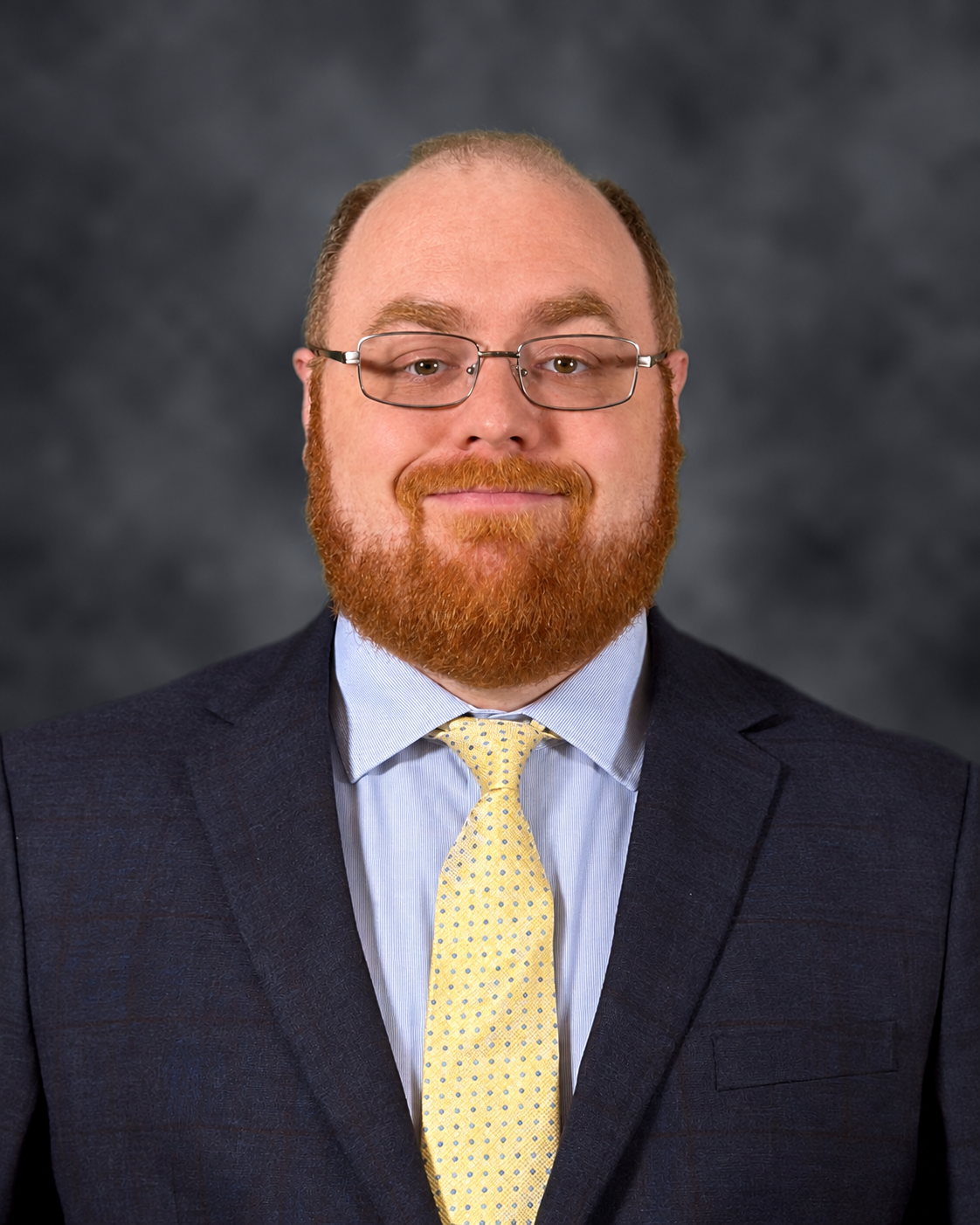 Headshot of a man with glasses, red beard, wearing a navy suit, light blue shirt, and yellow dotted tie, against a gray background.