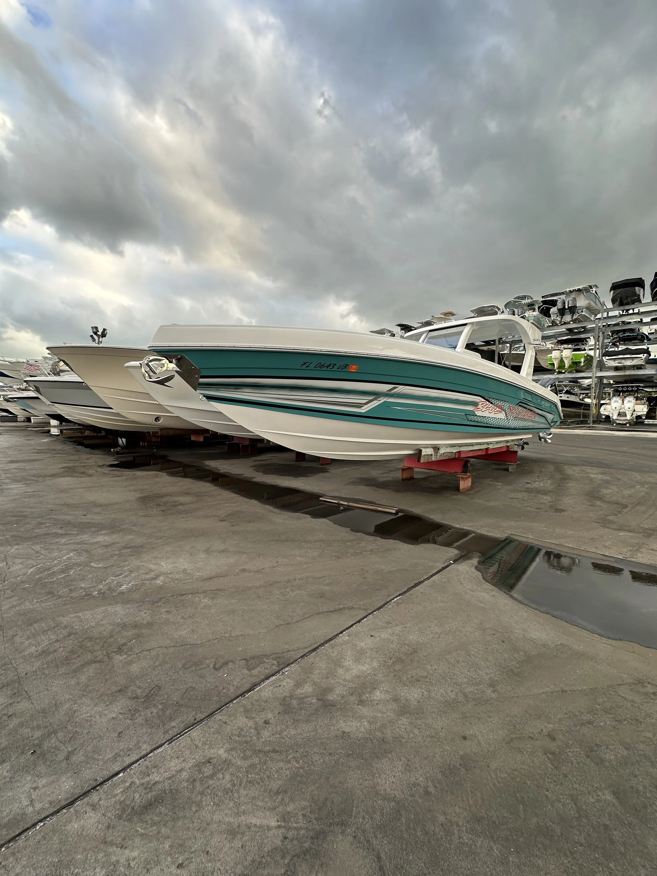 Teal-and-white speedboat displayed on a trailer at a marina, with stacked boats under a cloudy sky.
