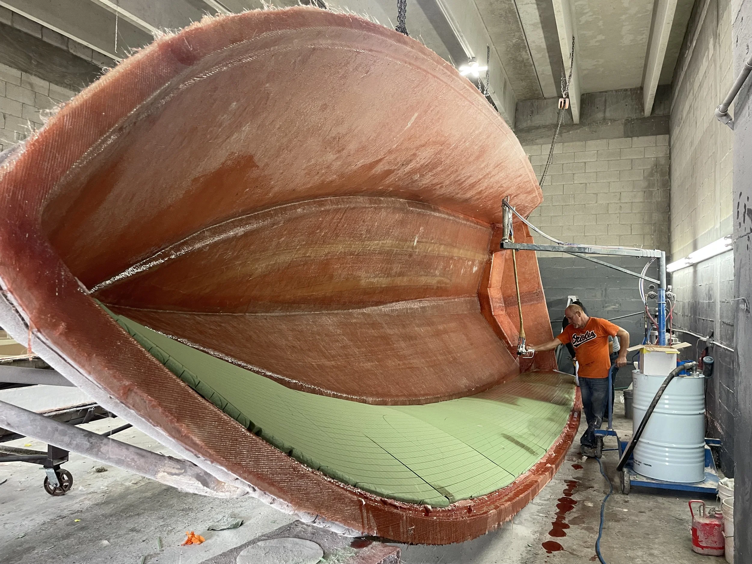 Large fiberglass boat hull under construction in a workshop, with workers and equipment nearby