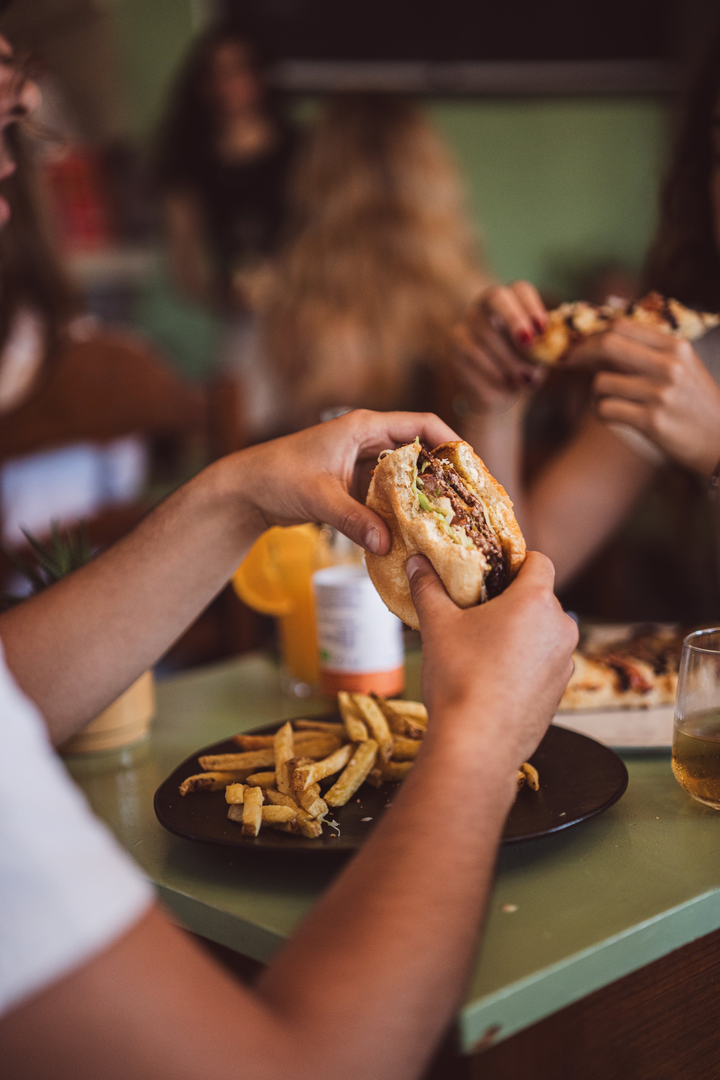Two people are holding burgers in a dining setting, with one person having a plate of French fries on the table nearby, and a glass of a yellow beverage.