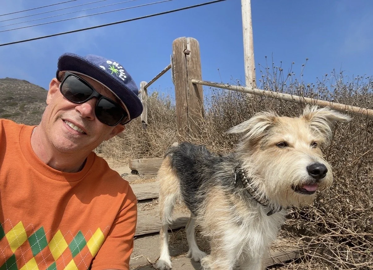 A man wearing sunglasses, a blue cap, and an orange shirt taking a selfie outdoors next to a small scruffy dog with light-colored fur and a darker patch on its back, with a dry, brown hillside and a wooden railing in the background.
