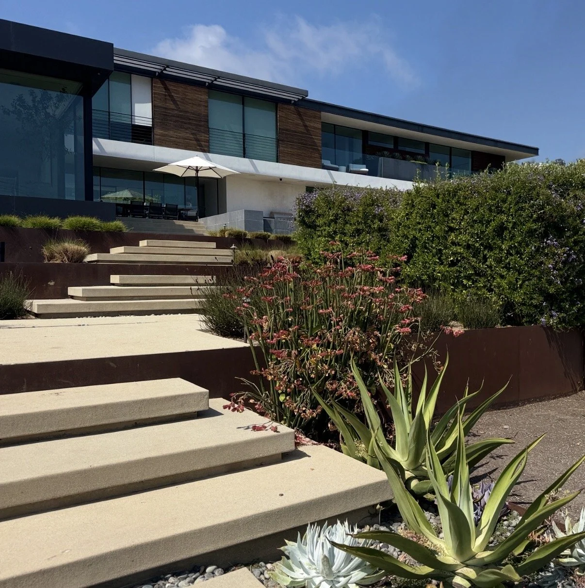 Modern house with large glass windows, a white and gray exterior, and an outdoor patio with an umbrella. The house is situated on a landscaped hillside with stairs, flowering plants, and lush greenery in the foreground. Clear blue sky overhead.
