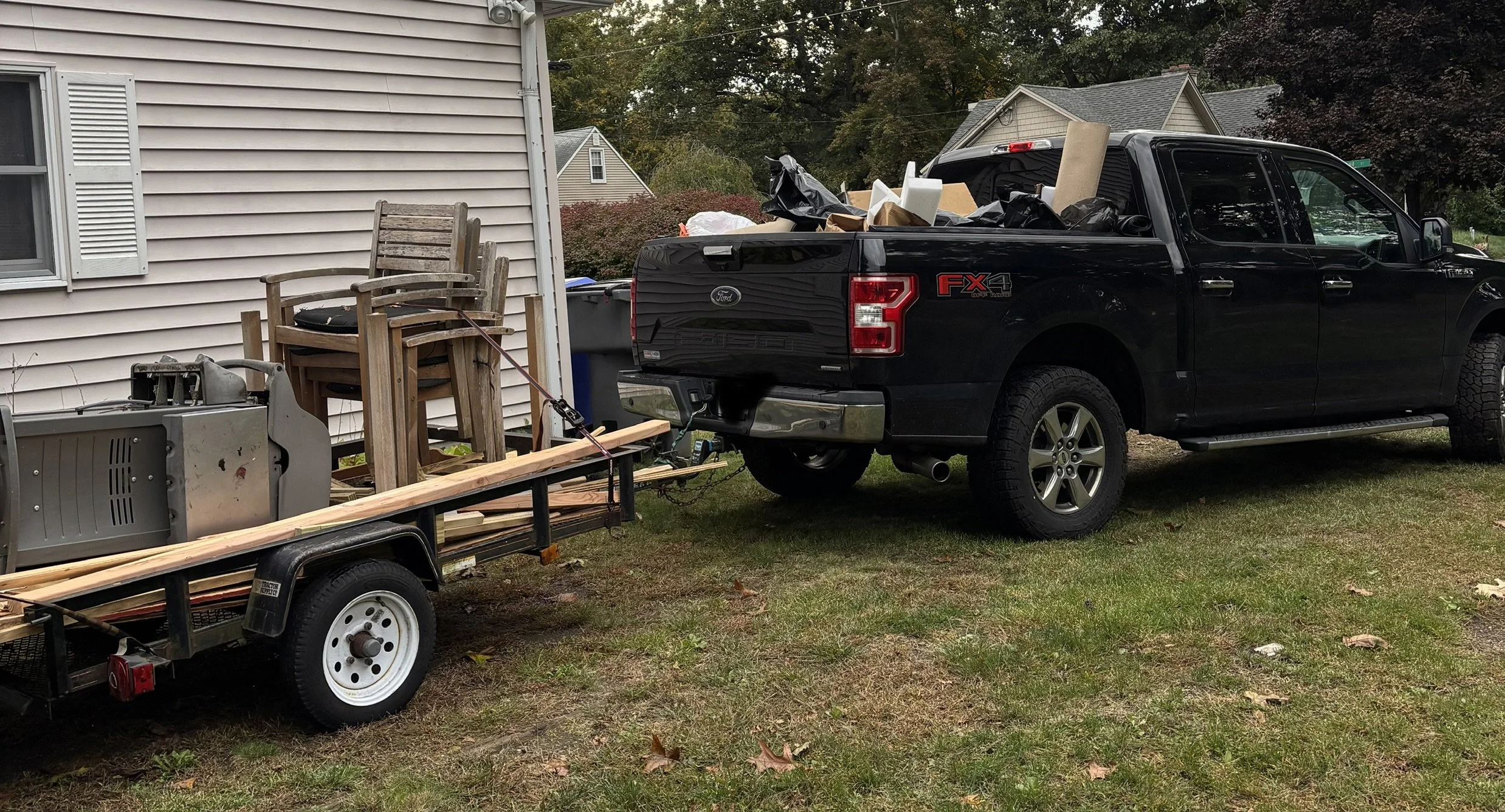 A black Ford pickup truck is parked on a lawn next to a house. The truck's bed is filled with various items including boxes, bags, and a piece of carpet. In front of the truck, there is a trailer loaded with old wooden furniture, a microwave, and a wooden plank.
