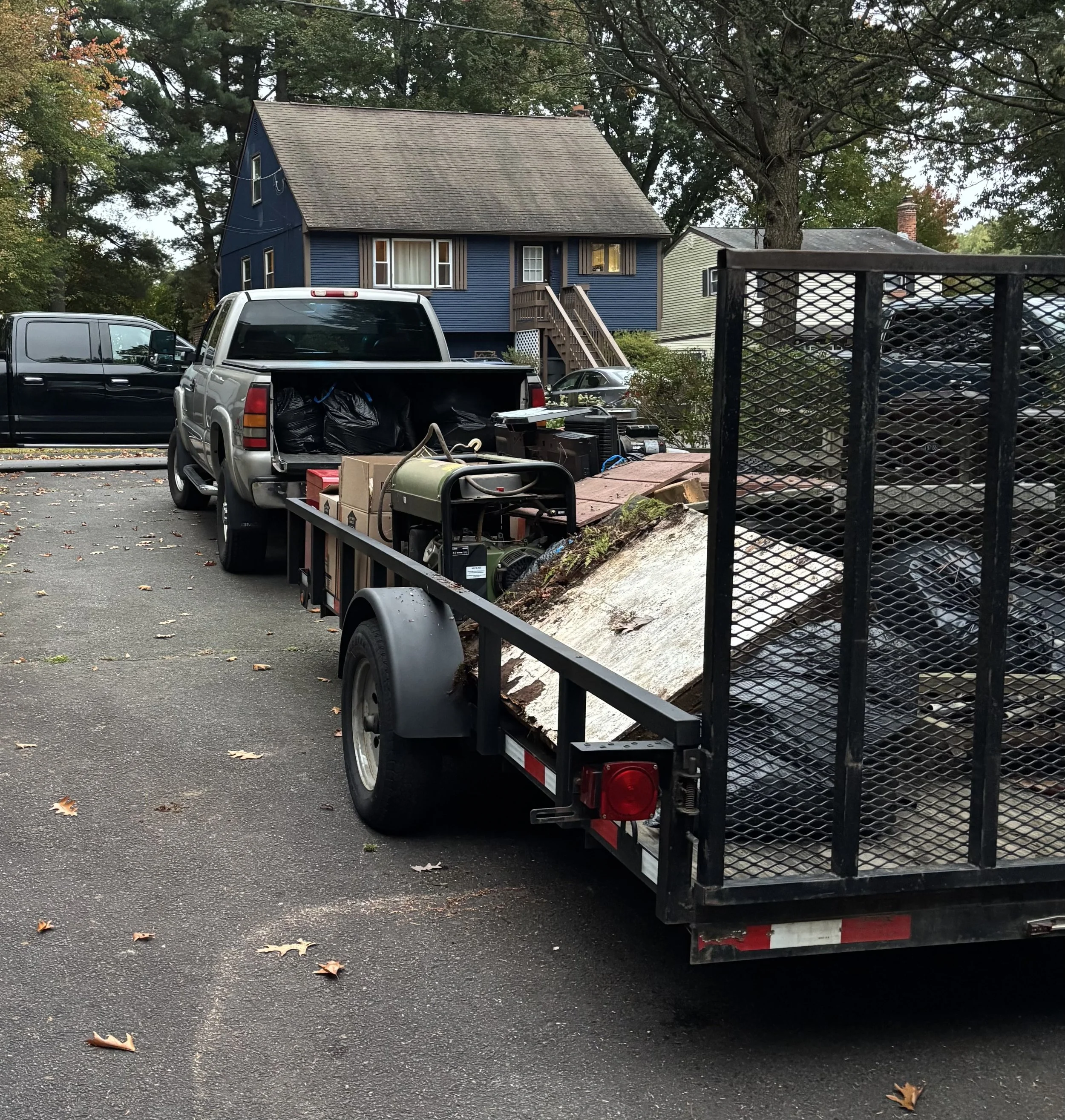 A yard with a blue house, several parked cars, a trailer filled with boxes and bags, and various tools and supplies on the ground.