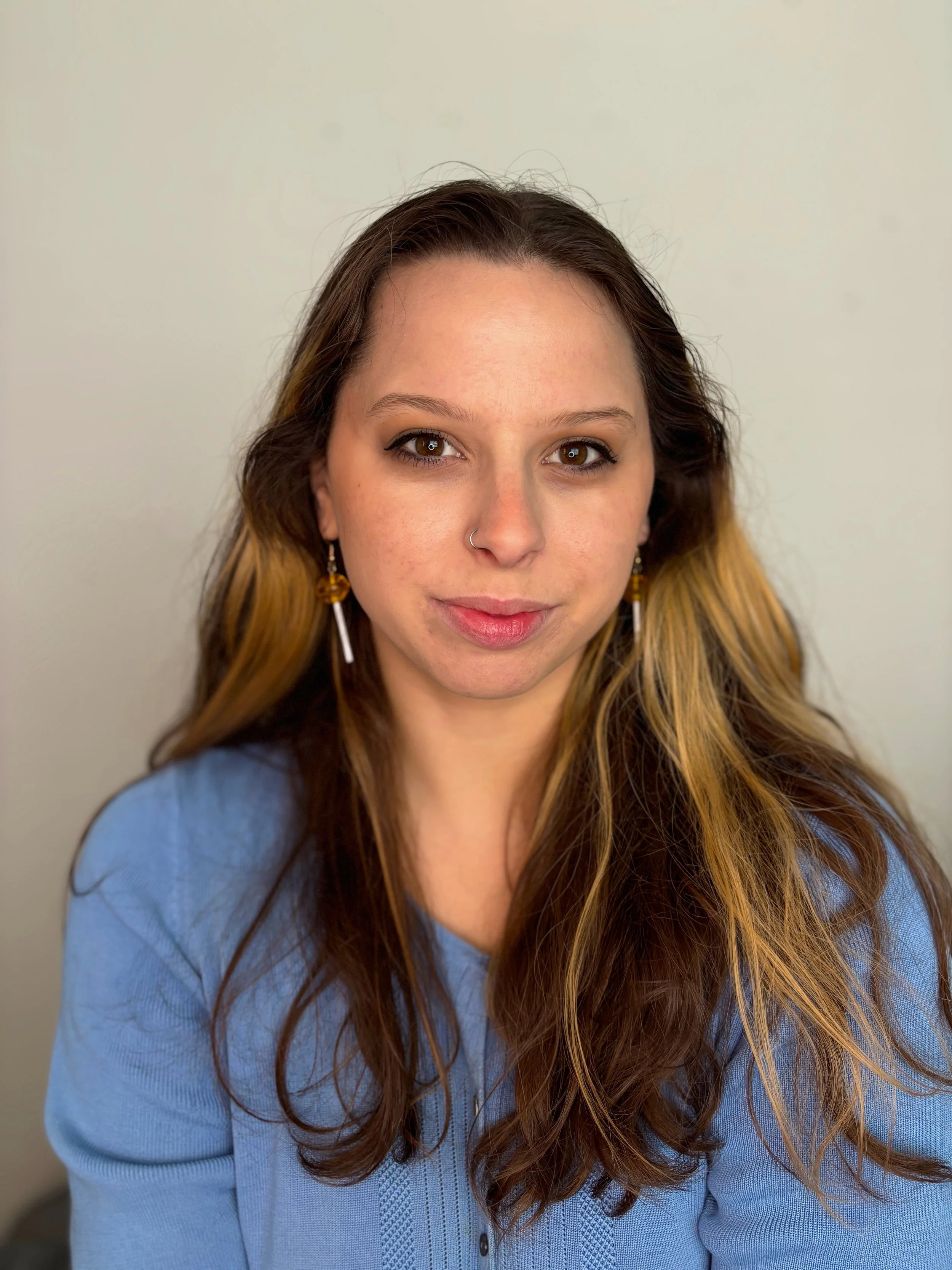 Close-up portrait of a woman with long brown hair, winged eyeliner, nose ring, and dangling earrings wearing a blue shirt