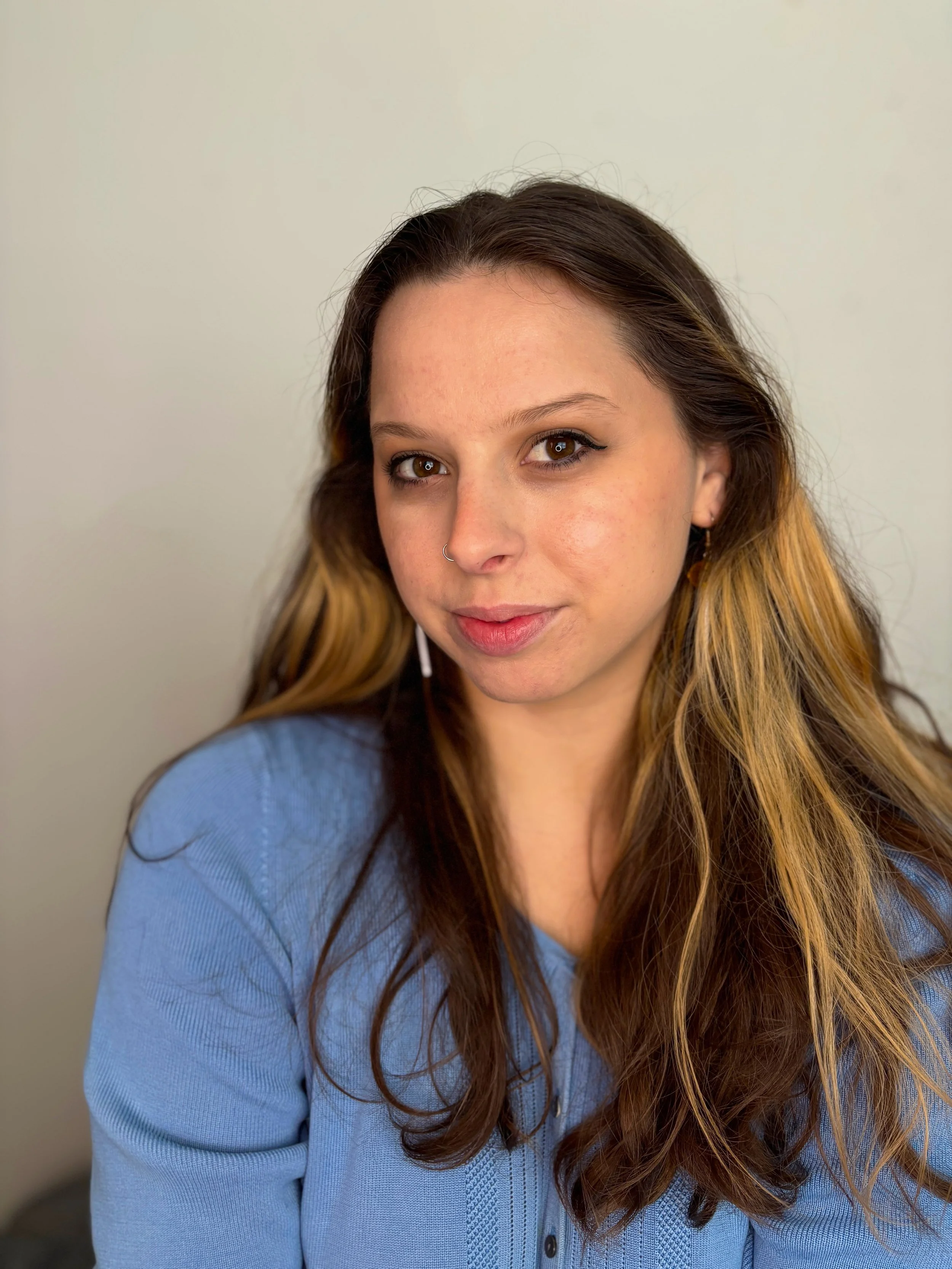 Close-up portrait of a woman with long brown hair, winged eyeliner, brown eyes, and a nose ring wearing a blue sweater