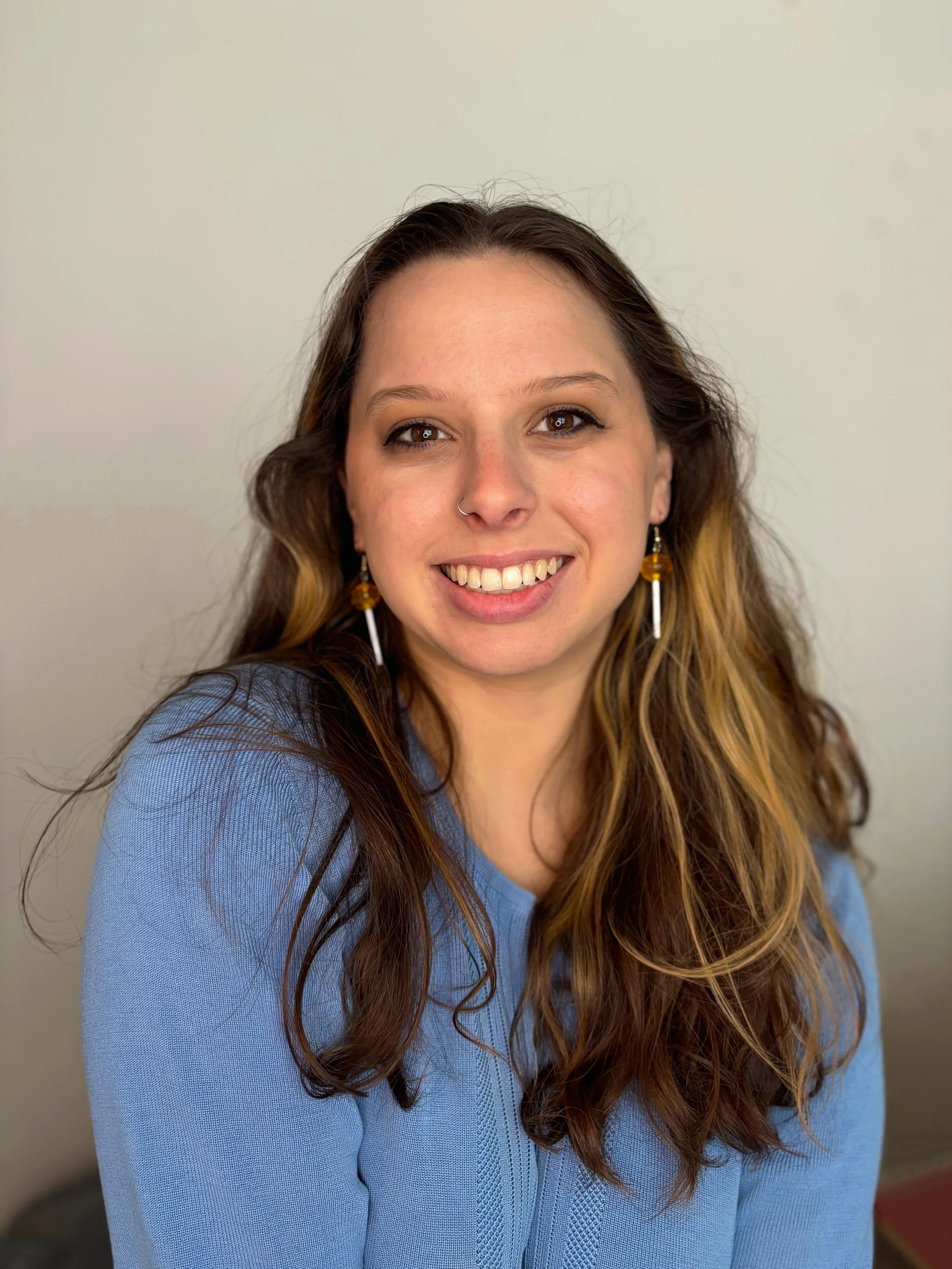 Smiling woman with long brown hair, hoop nose ring, and dangling earrings wearing a blue sweater