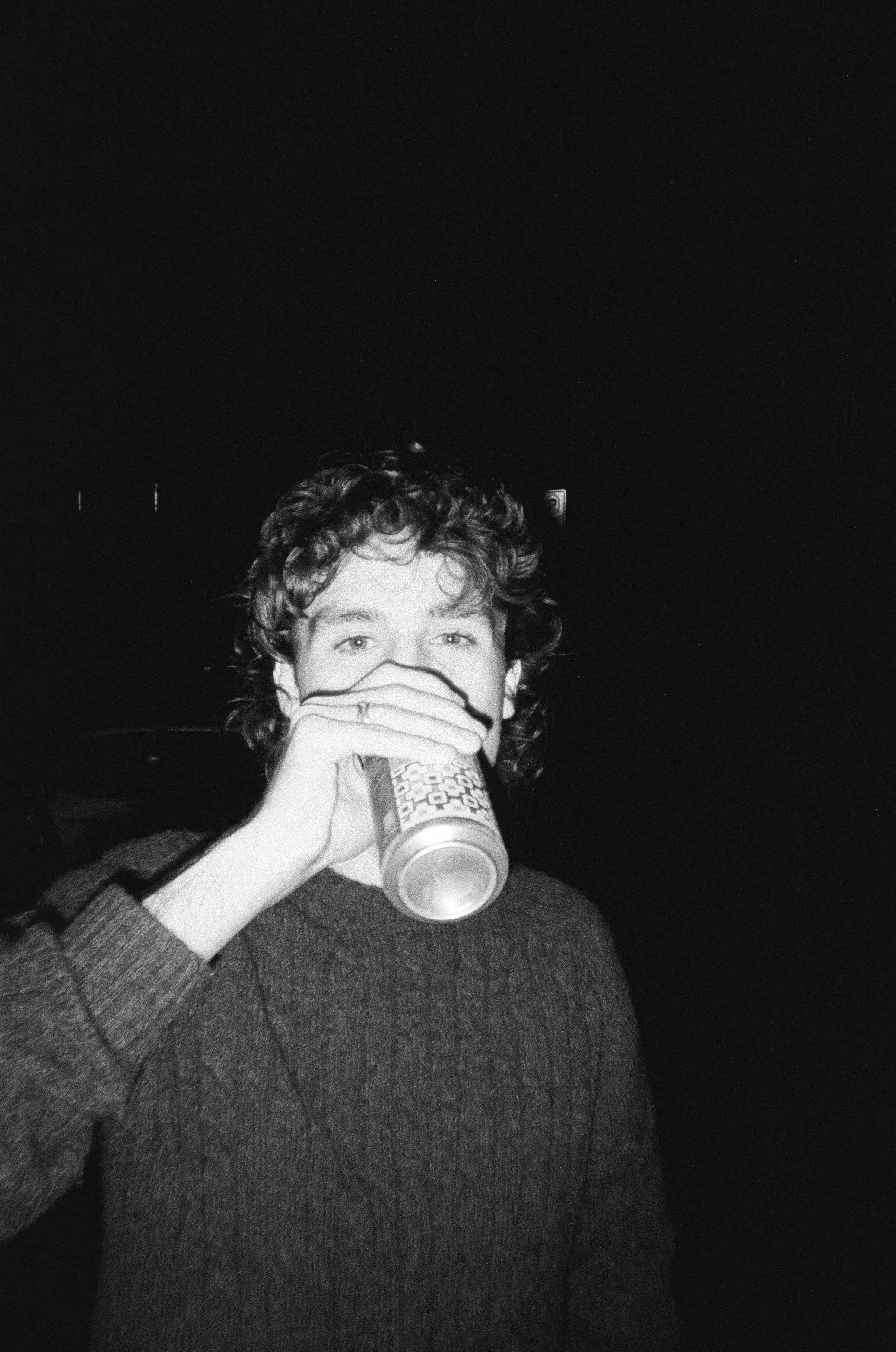 A person with curly hair drinking from a can, wearing a dark sweater, against a dark background.