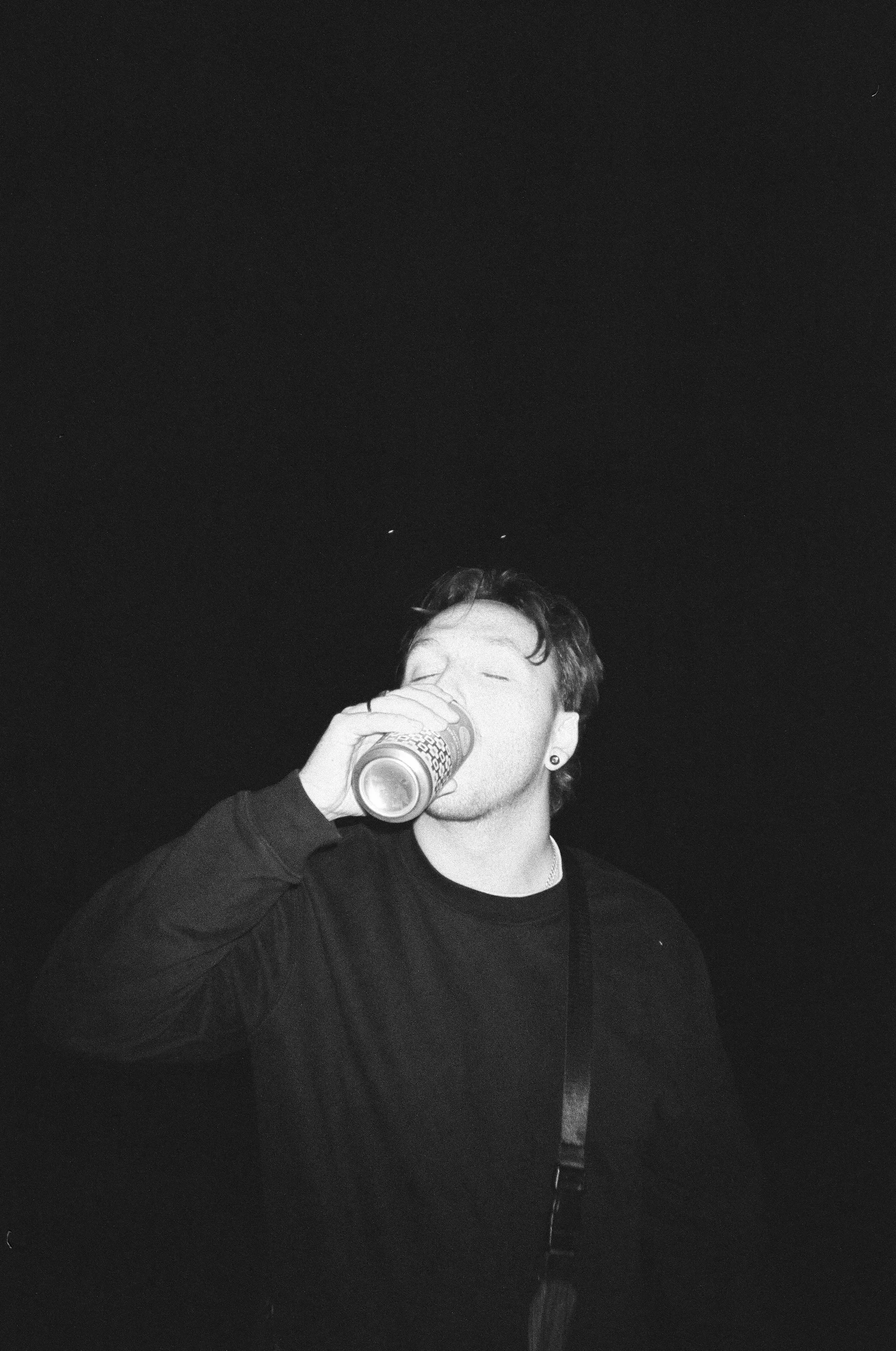 A young man with short, wavy hair drinking from a can in a dark setting.