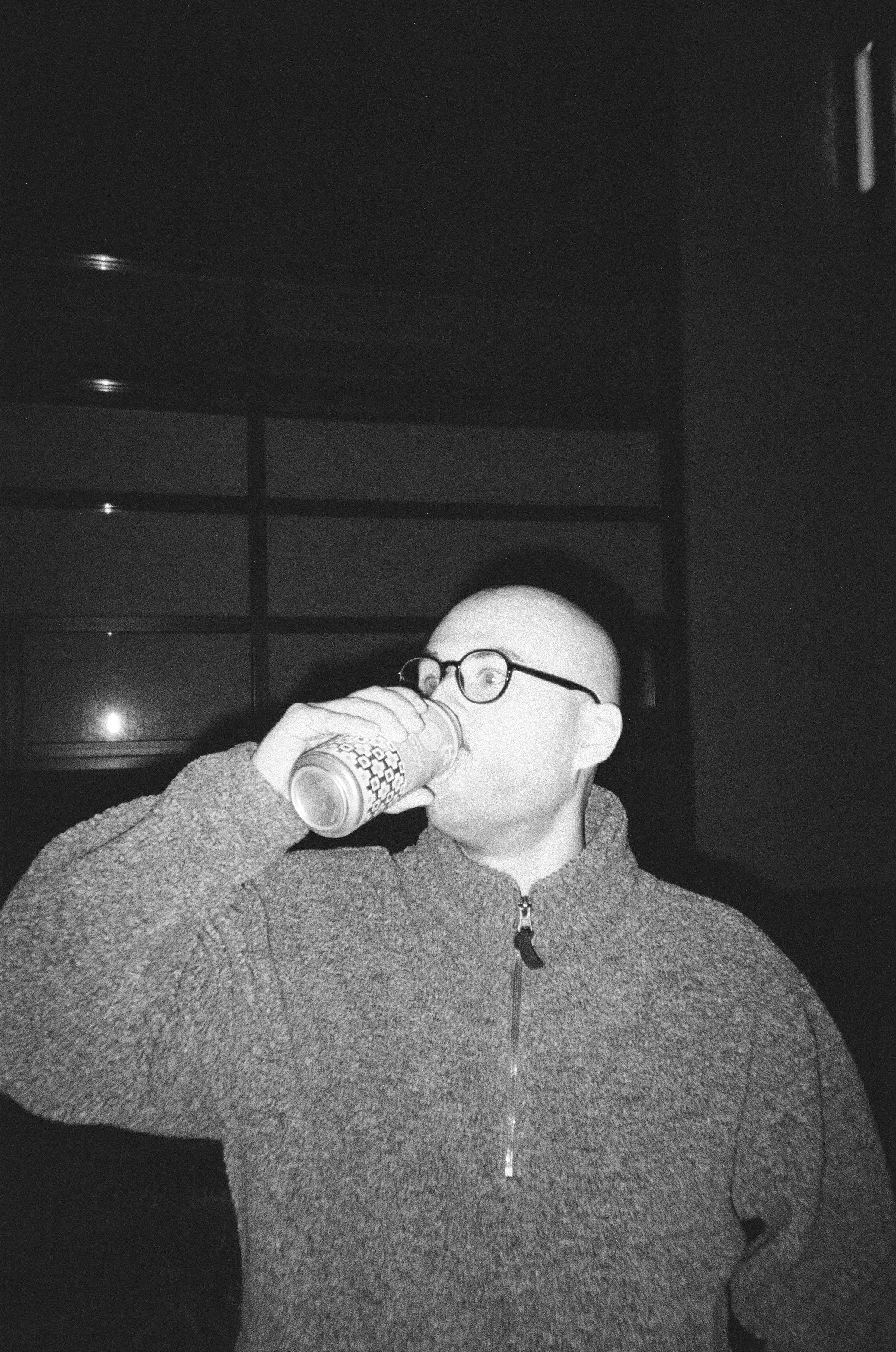 A person with glasses and a hoodie drinking from a cup in a dimly lit room.