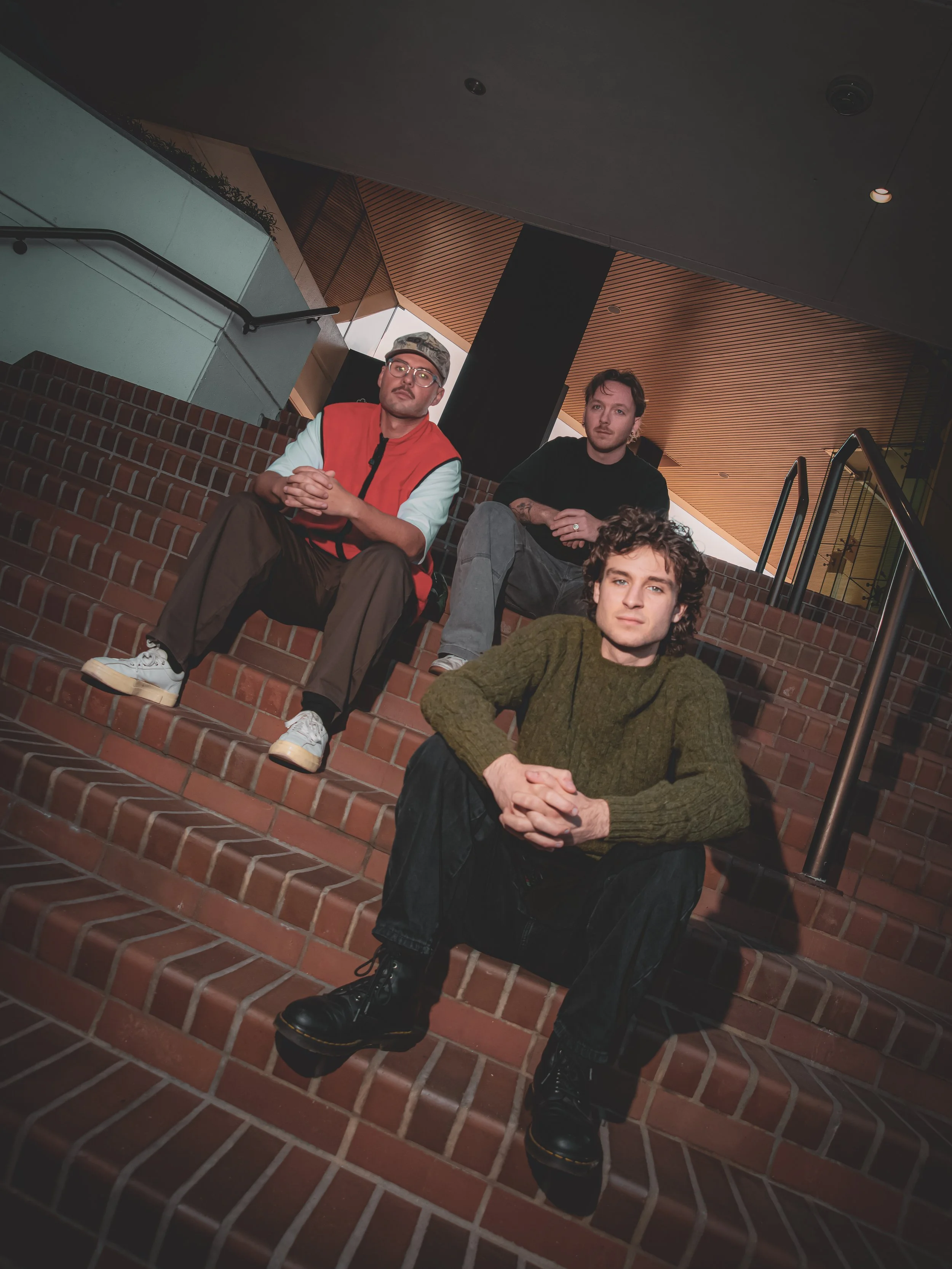 Three young men sitting on brick stairs in an indoor setting, with a modern architectural background featuring wood and black paneling.