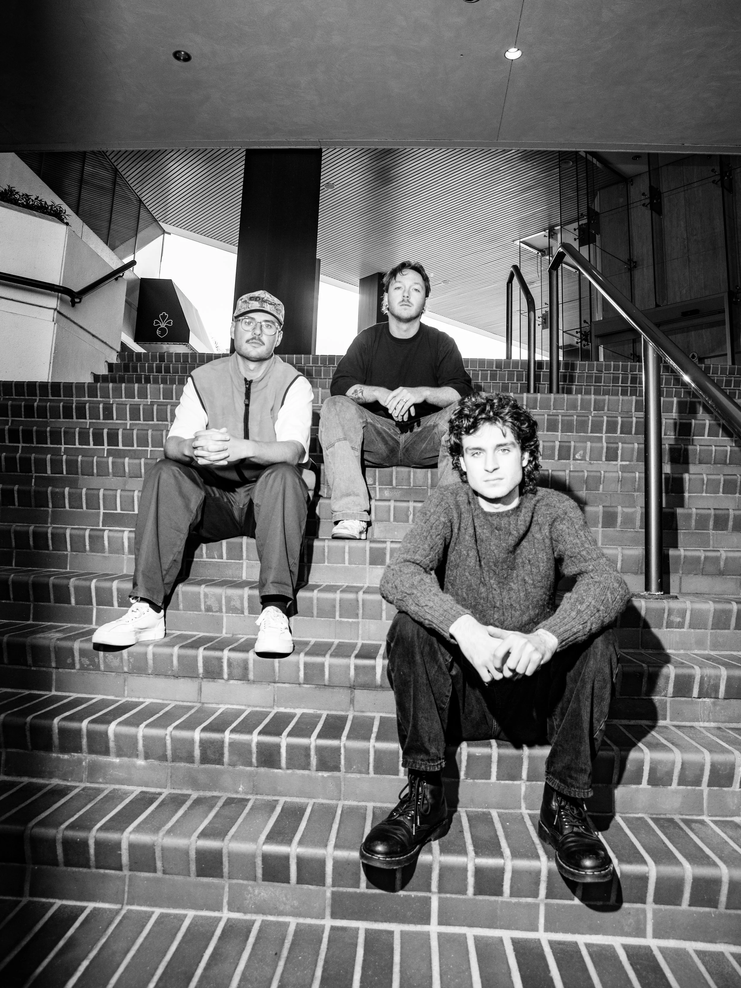 Four young men sitting on brick stairs inside a modern building with a high ceiling and metal railings.