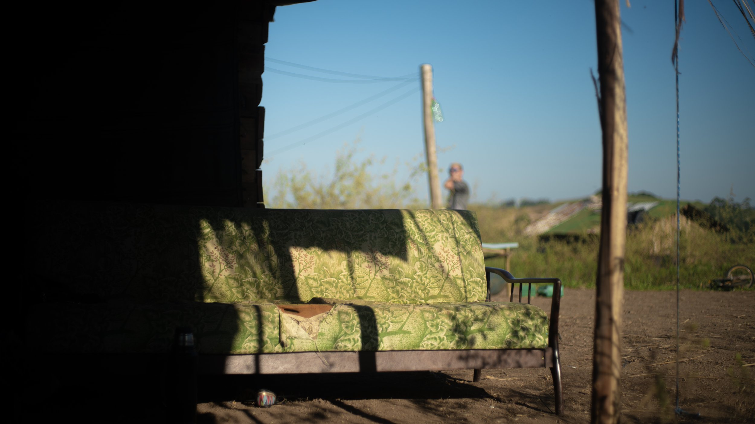A green patterned sofa, seen through a dark doorway, outdoors with a person in the background near a utility pole and power lines.