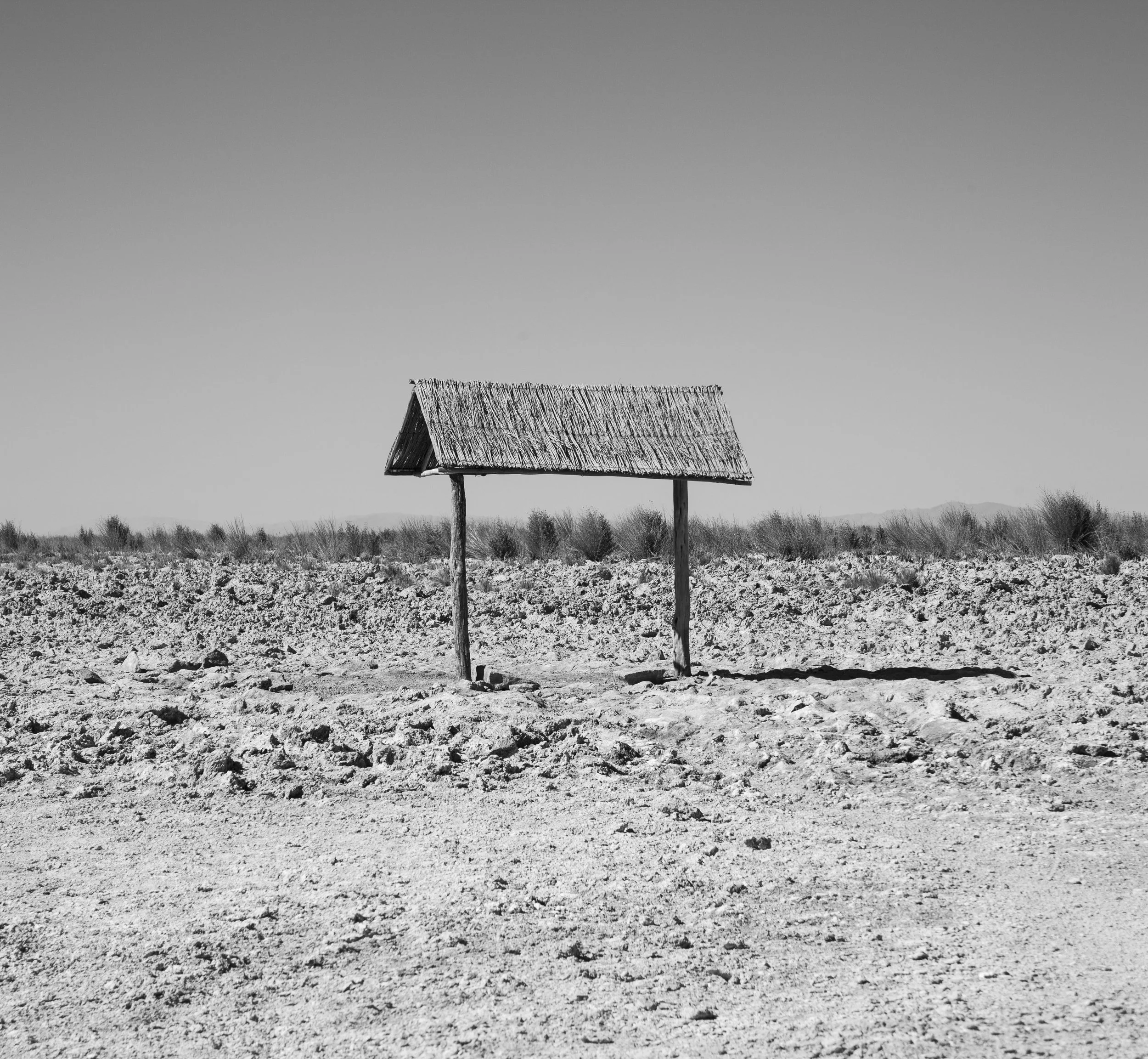 A solitary, weathered wooden structure with a thatched roof stands in a barren, arid landscape under a vast, empty sky.