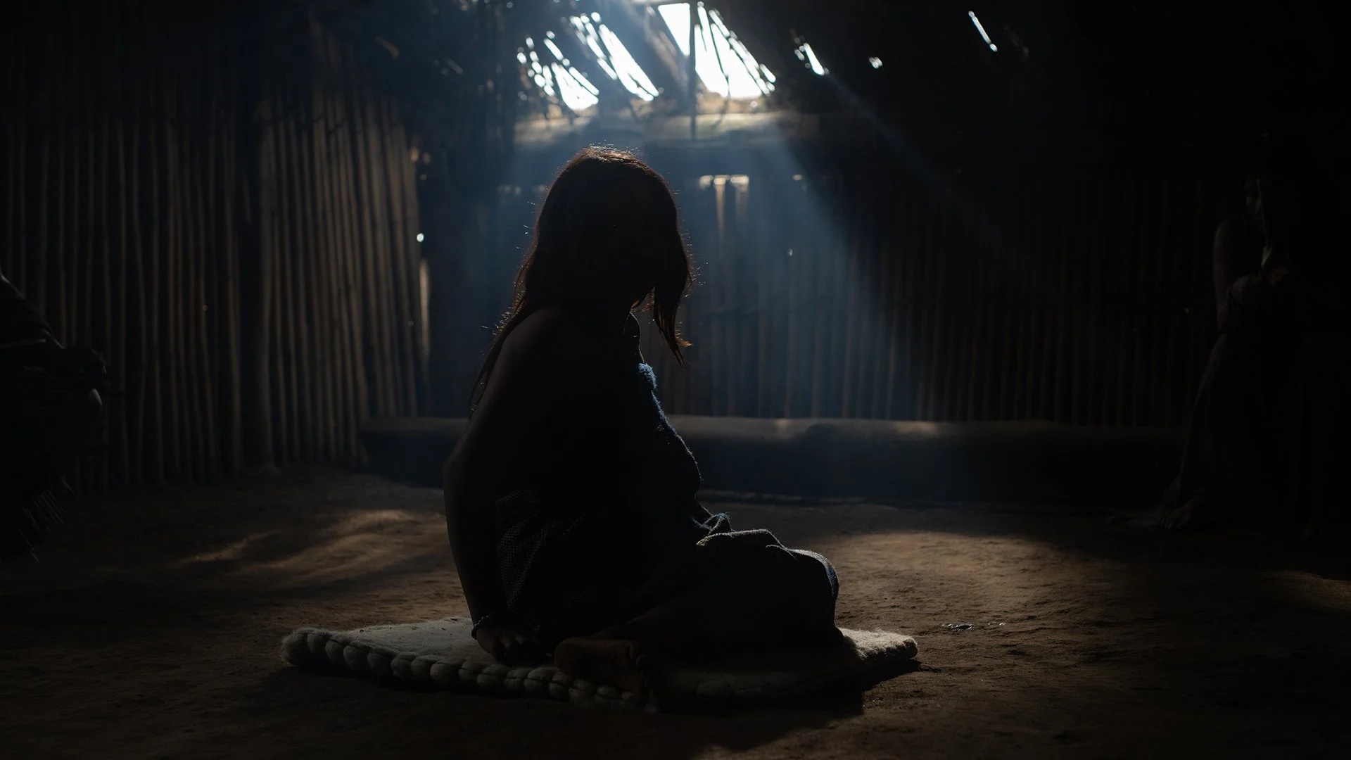 A silhouette of a person sitting on a mat inside a dimly lit hut, with sunlight streaming through the roof holes.