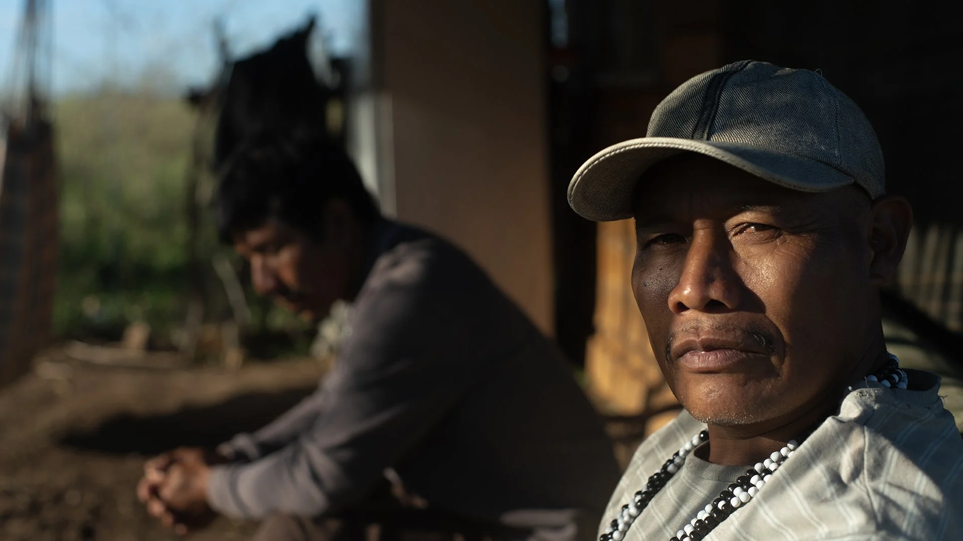 Two people sitting outdoors, the man in the foreground wears a gray cap and beaded necklace, while the woman in the background wears glasses. They are in a rural setting with blurred greenery and structures.