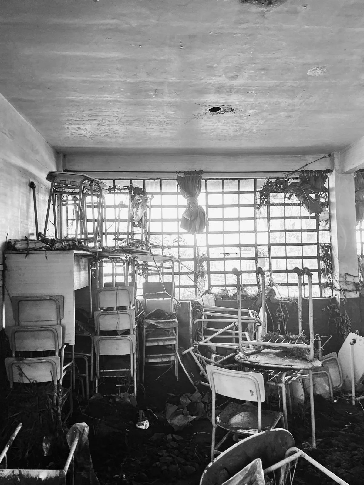 A room filled with charred furniture, including chairs stacked on top of each other, tables, and a hospital bed, with broken windows and debris on the floor.