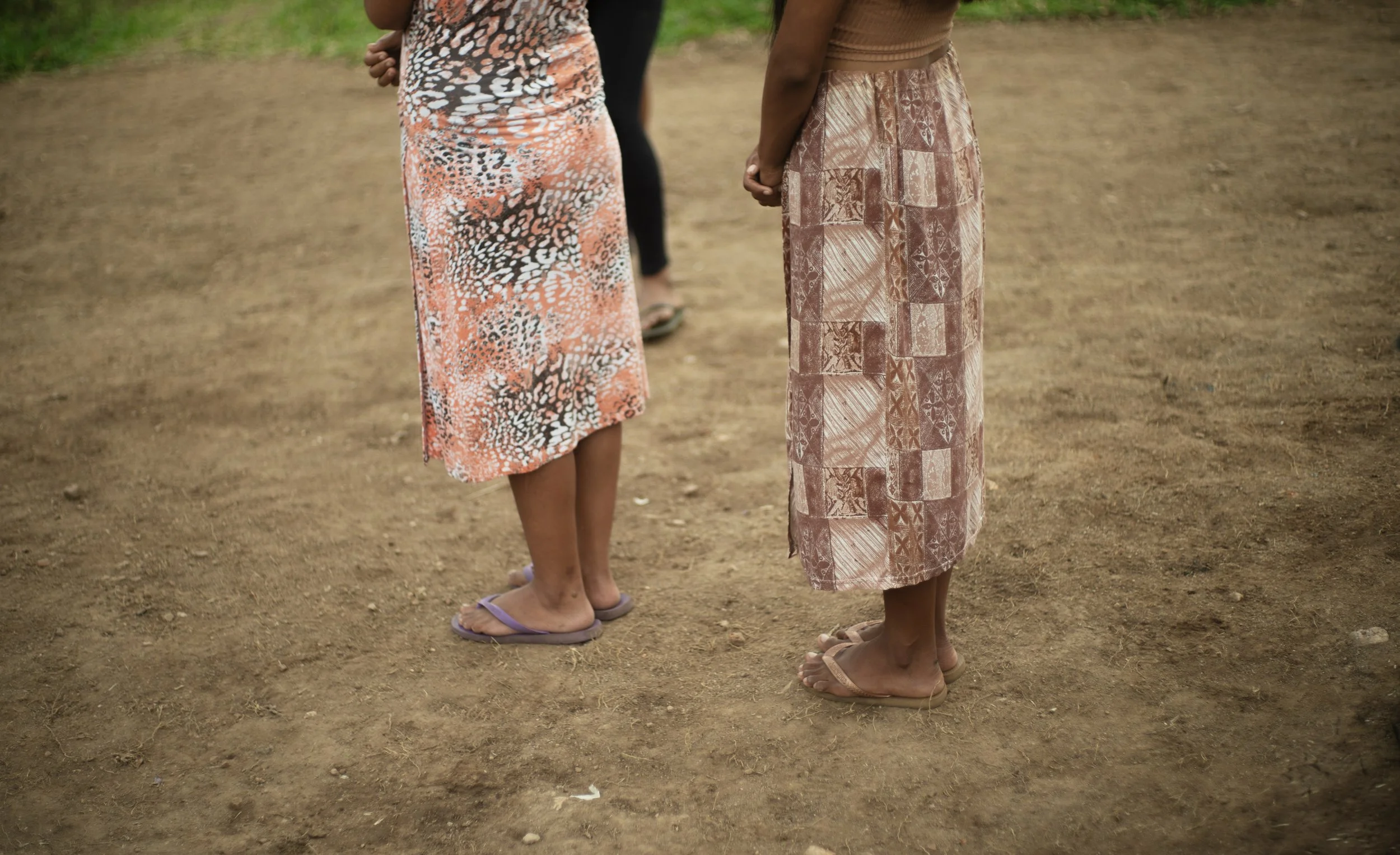 Two girls standing on dirt ground, holding hands behind their backs, wearing colorful dresses and sandals. They are outdoors, with greenery in the background.