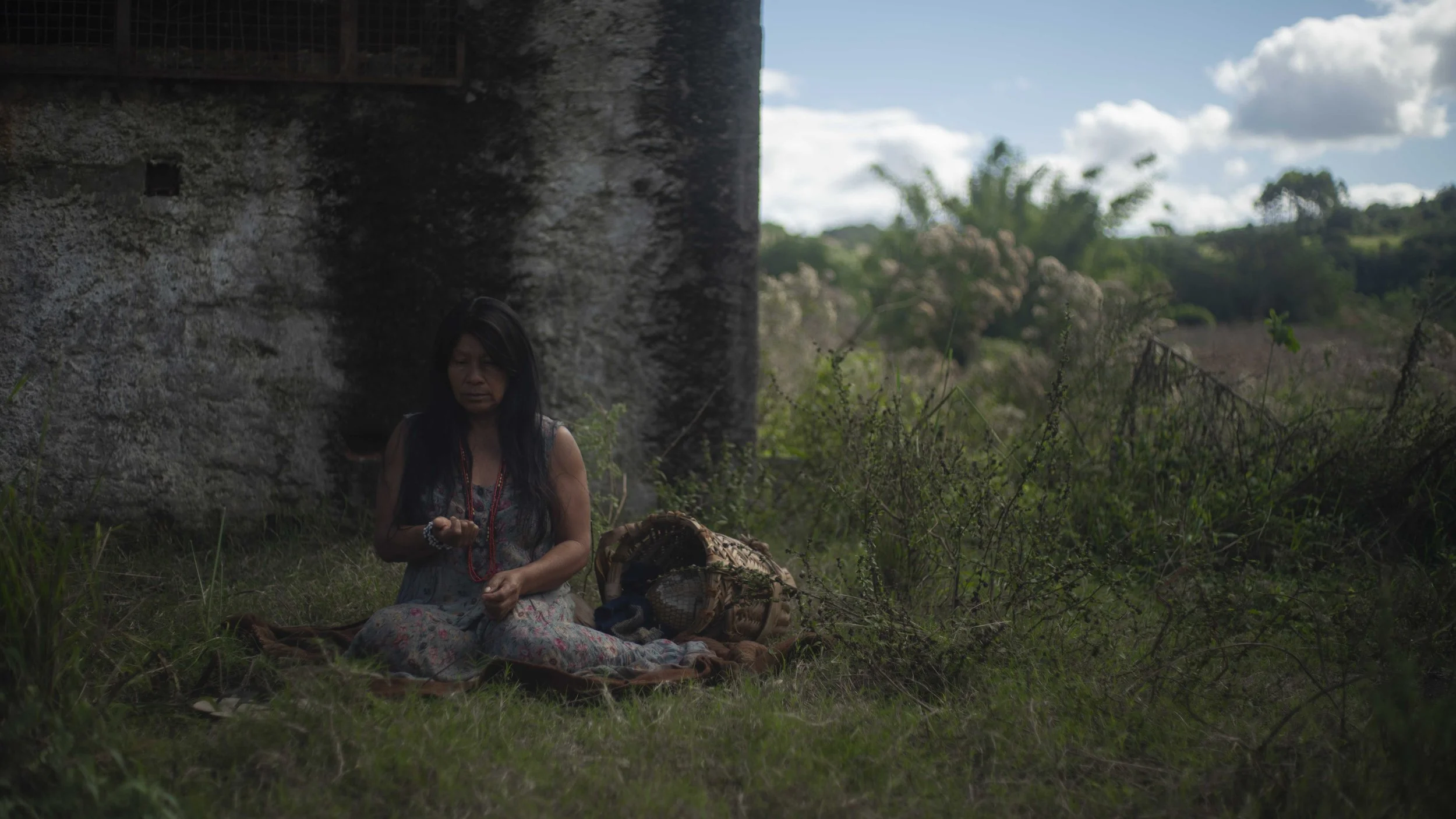 A woman sitting outdoors under a stone bridge, surrounded by grass and plants, with a basket nearby and a scenic background of trees and sky.