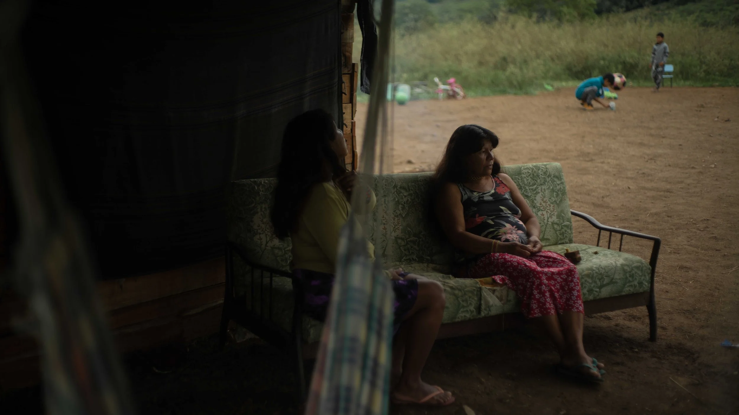 Two women sitting on a patterned green sofa inside a dimly lit structure, looking out into a dirt yard where two children and a young man are playing outdoors. The scene depicts a rural setting with grassy areas and outdoor toys.