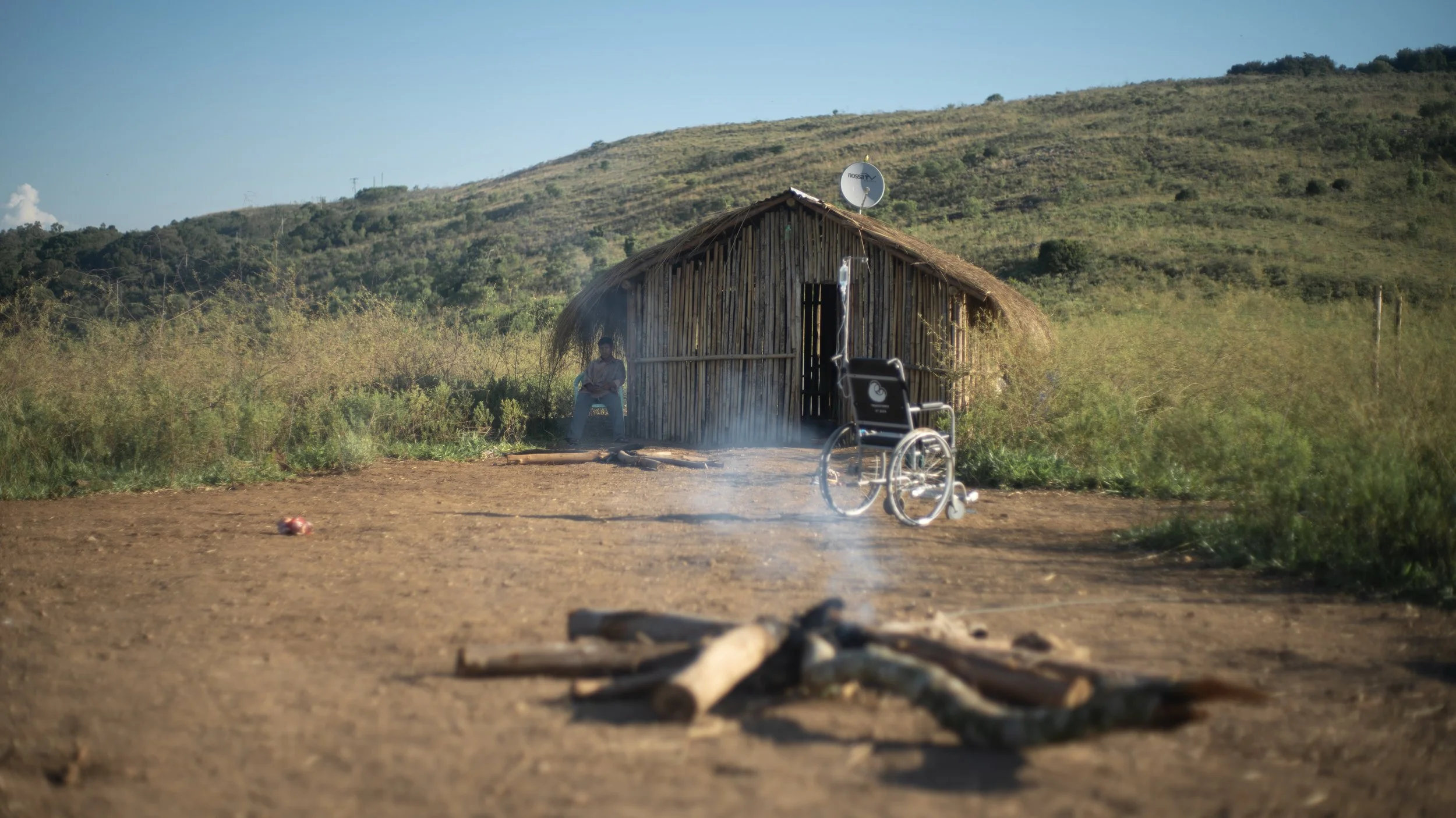 A wheelchair outside a rustic wooden hut in a rural landscape with hilly terrain, smoke on the ground, and a man sitting on a chair near the hut.
