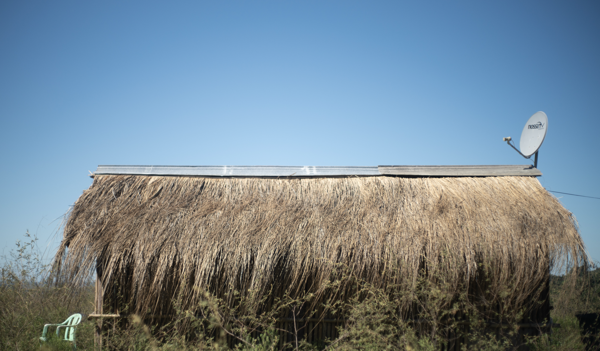 Thatched roof house with a satellite dish labeled "NOSSA TV" and a clear blue sky.