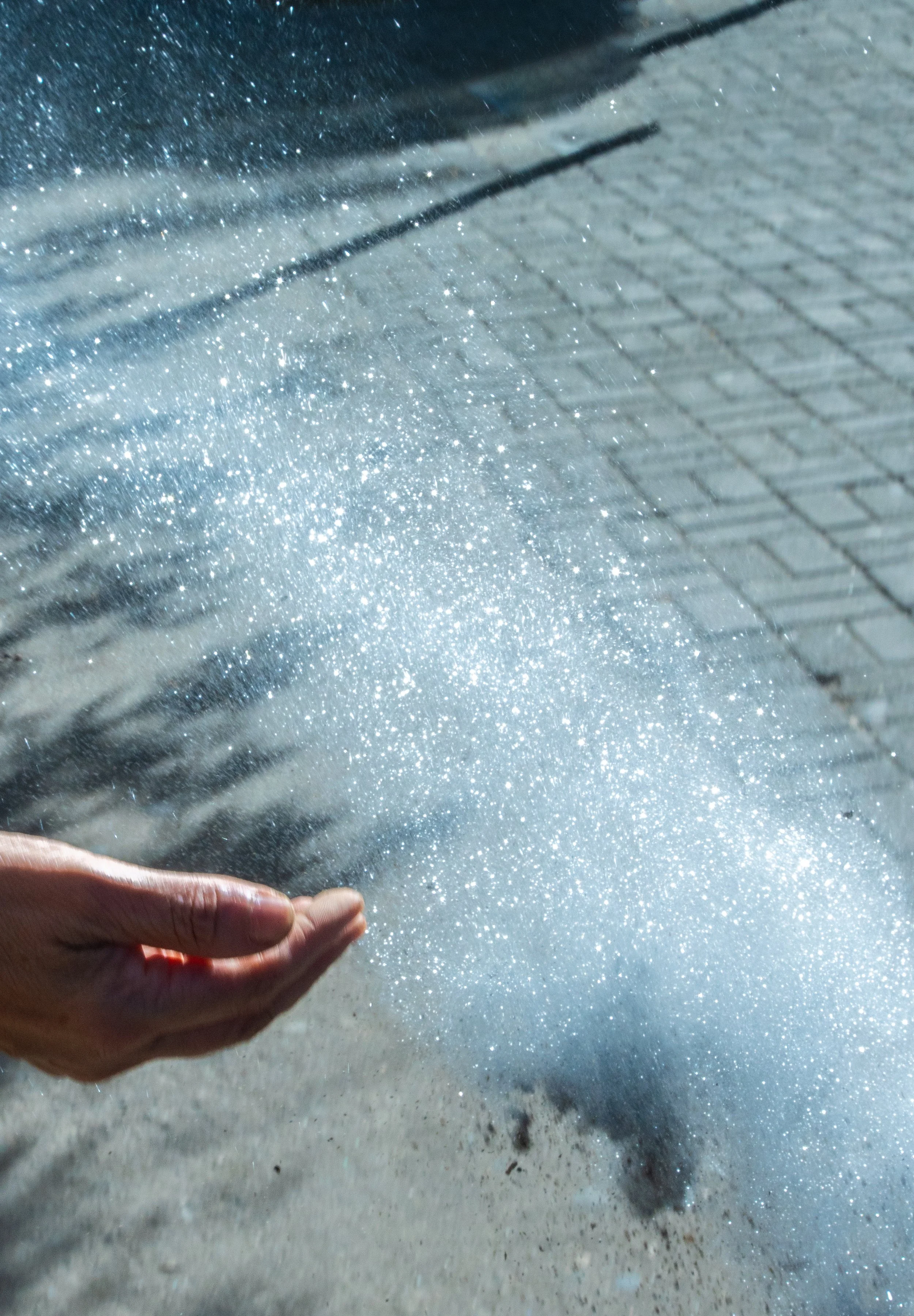 A person holding a handful of shimmering sand or glitter on a beach, with the sandy ground and a concrete or tiled surface in the background.