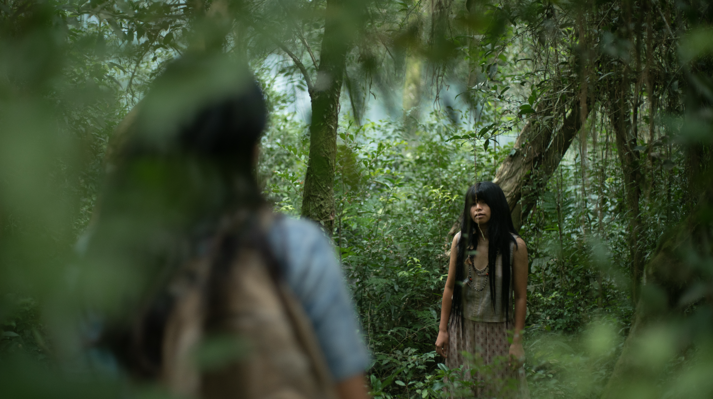 A woman with long dark hair standing in a lush green forest, wearing a dress and layered necklaces, with trees and foliage surrounding her.