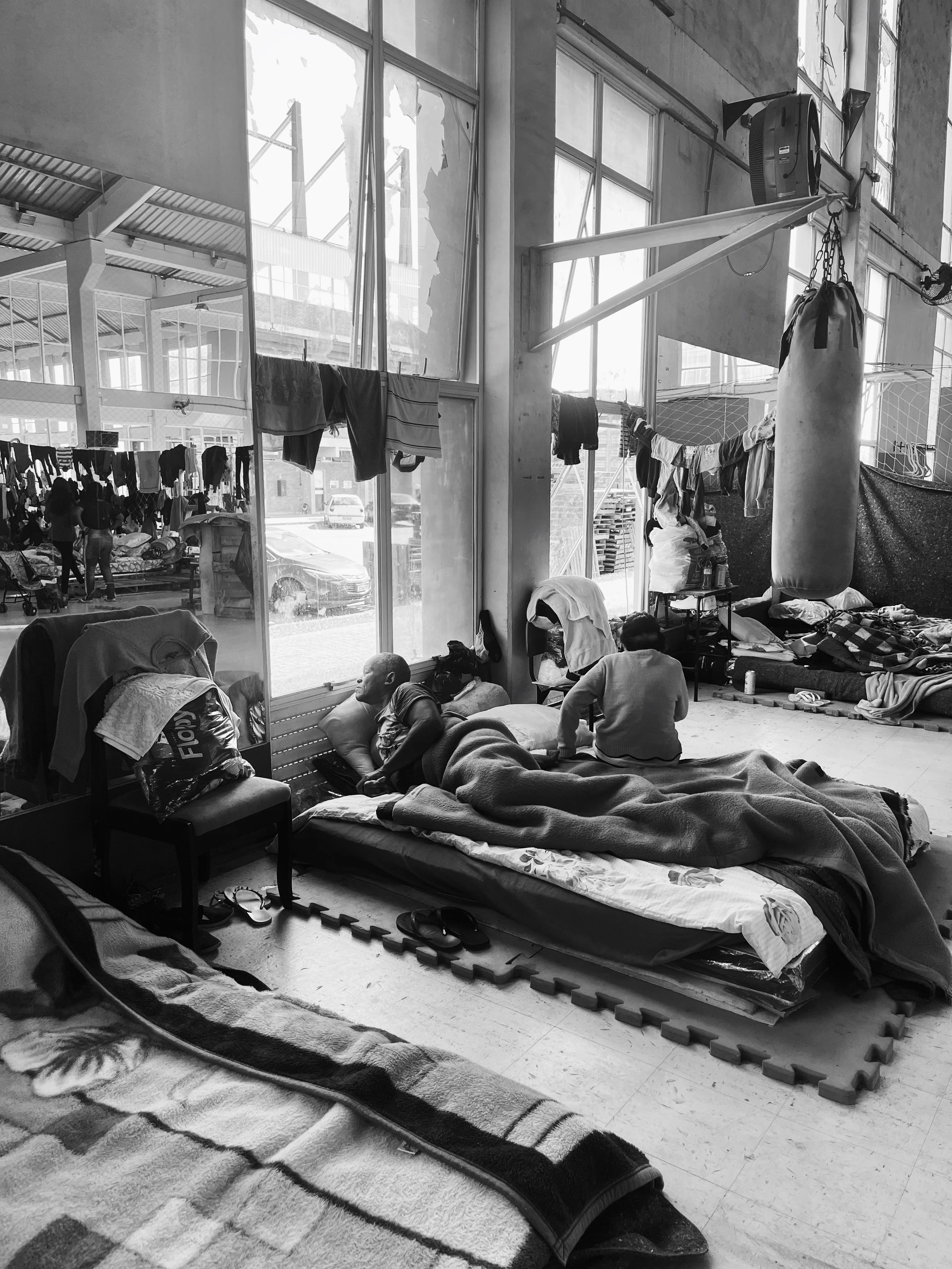People resting and sleeping inside a large, industrial-style building with large windows. Sleeping mats and blankets are laid out on the floor, with some individuals lying down and others sitting up. Laundry hangs from lines near the windows. There are chairs, a punching bag, and various personal belongings scattered around the space.