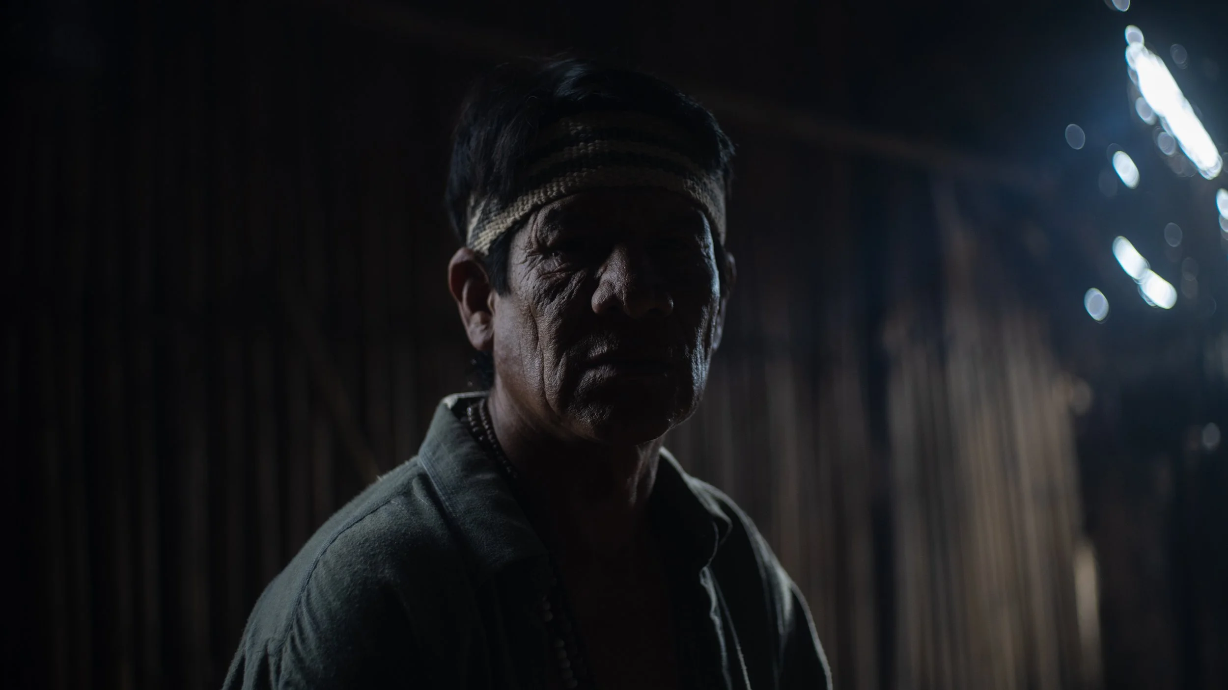 A man wearing a headband and a dark jacket, with a serious expression, standing in a dimly lit environment with streaks of light in the background.
