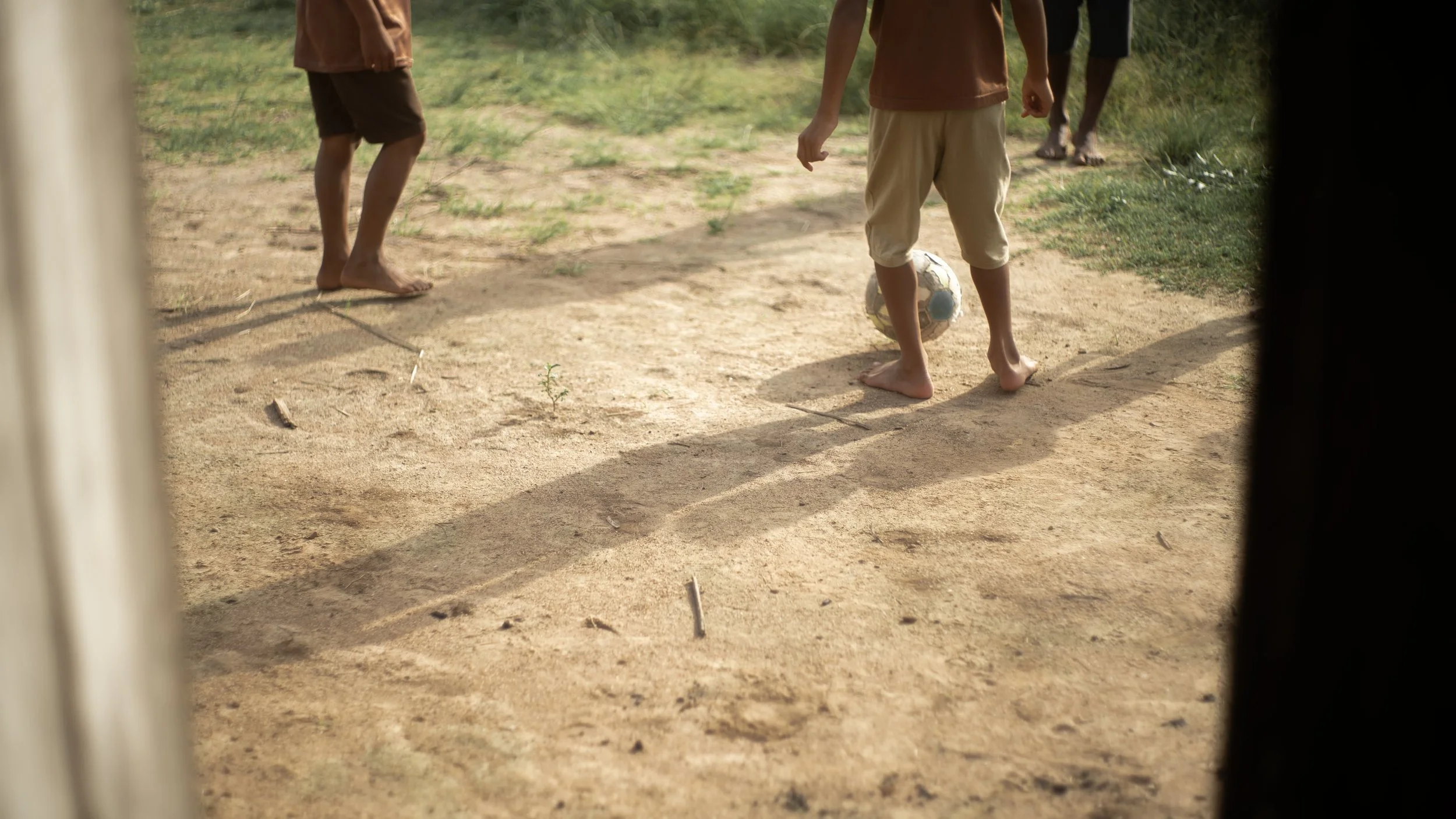 Children playing soccer on a dirt field outdoors, viewed through a window frame.