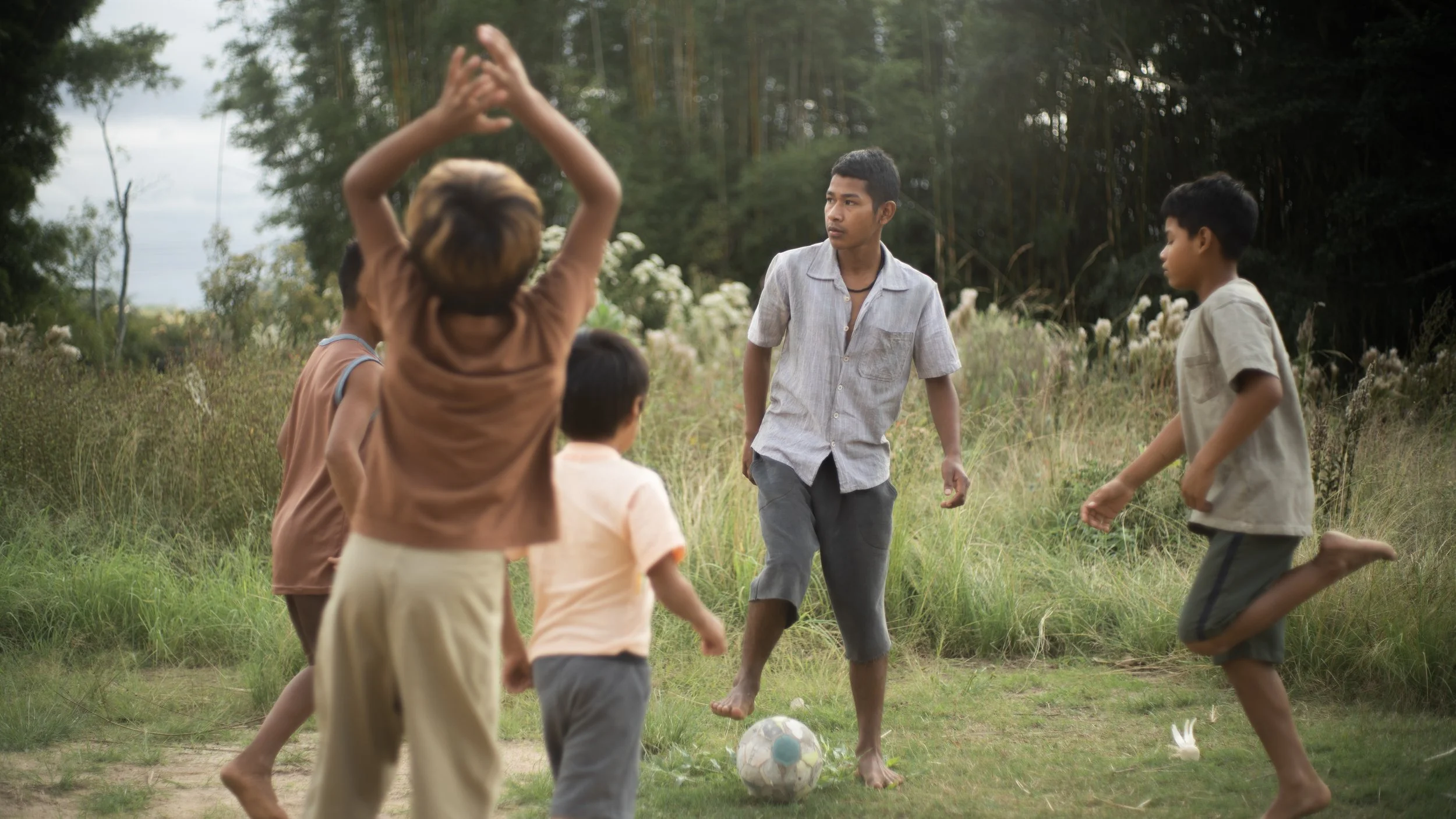 A group of children playing soccer outdoors in a grassy field surrounded by trees, with a young man in gray shorts and a light shirt standing among them.