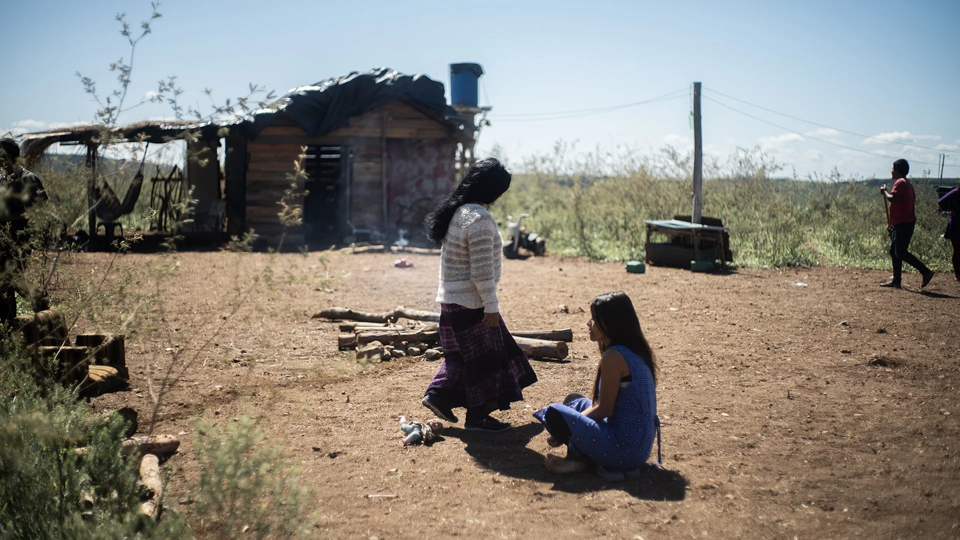 Two women sitting on the ground and talking outside a small wooden house, with one standing. Other people are in the background, some near a table and electric pole in a rural setting under a clear sky.