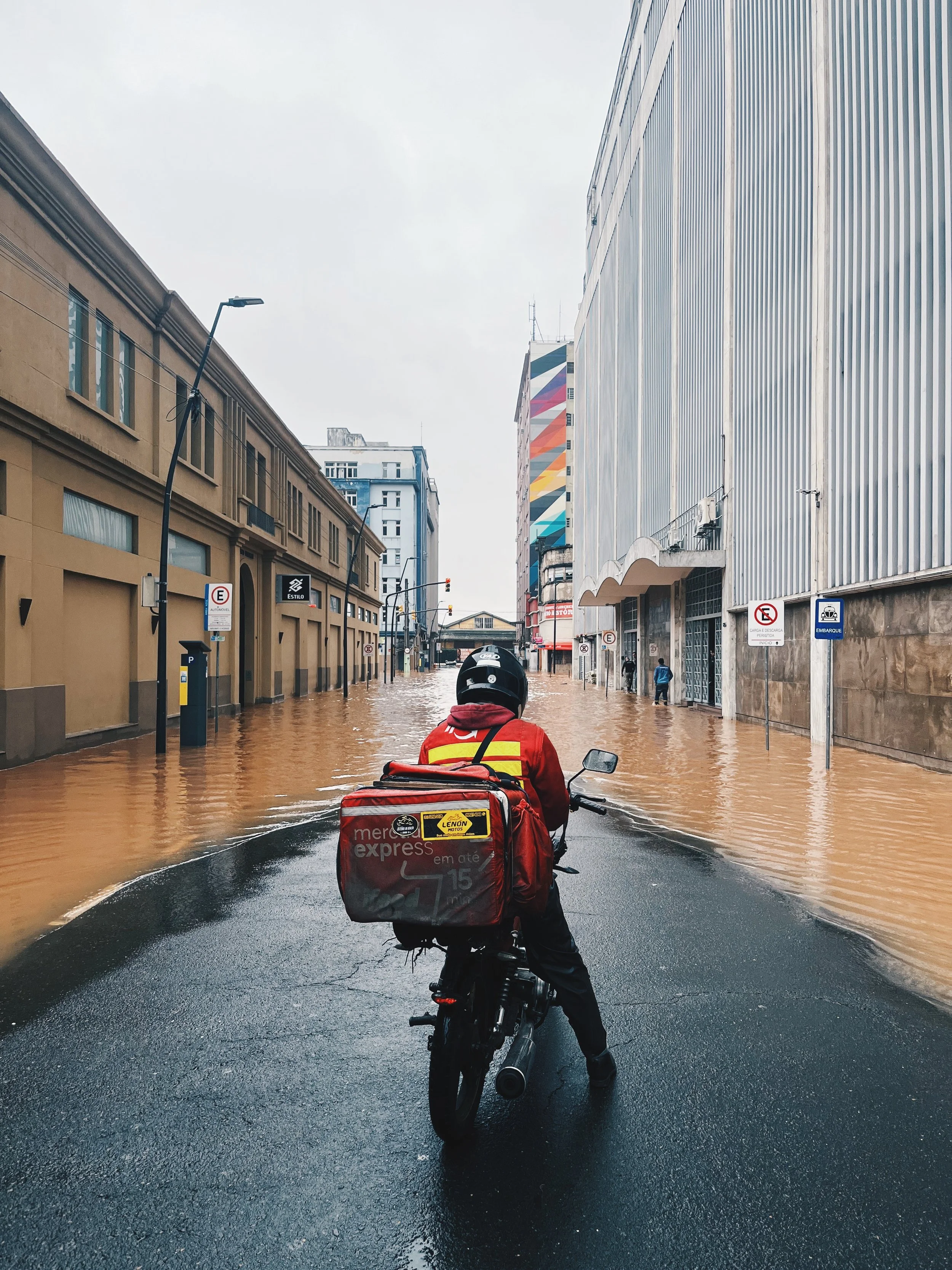 A person riding a motorcycle through a flooded city street during a rainstorm, with tall buildings on either side and water covering the road.