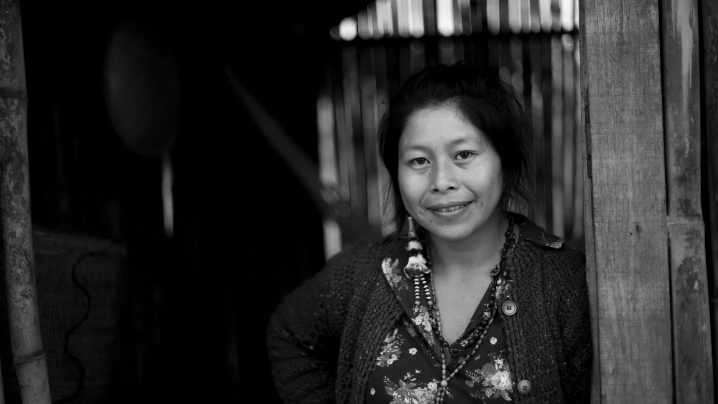 Black-and-white photo of a woman with short, dark hair smiling, standing outside near a wooden structure, with bookshelves in the background.