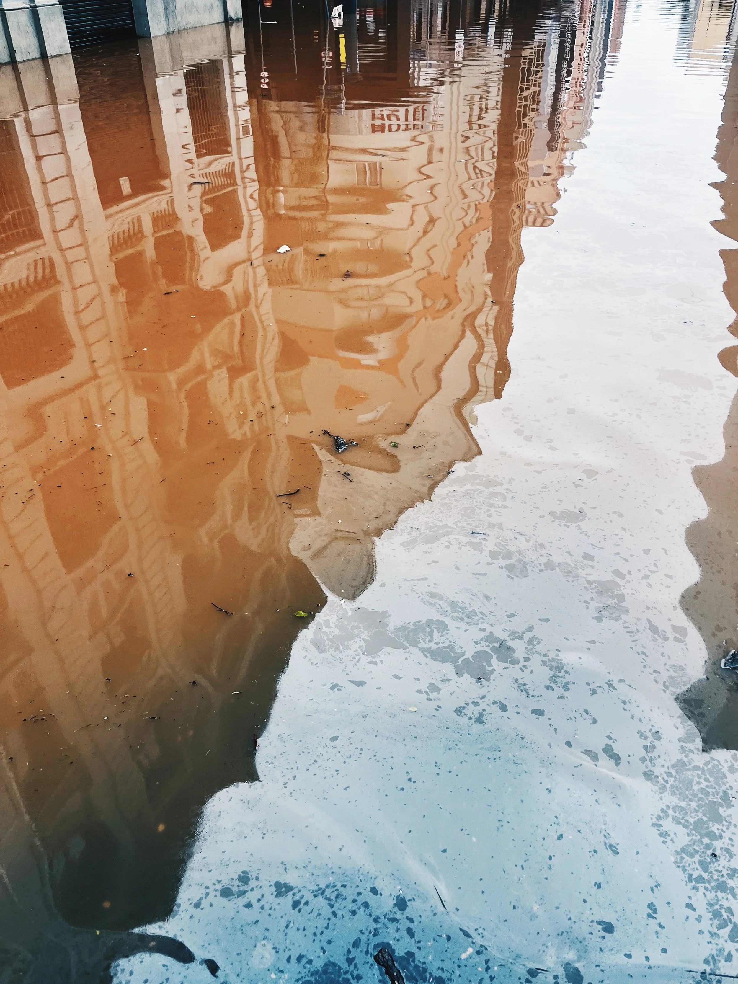 Reflected image of buildings and a street in a large puddle of muddy water on a city sidewalk.