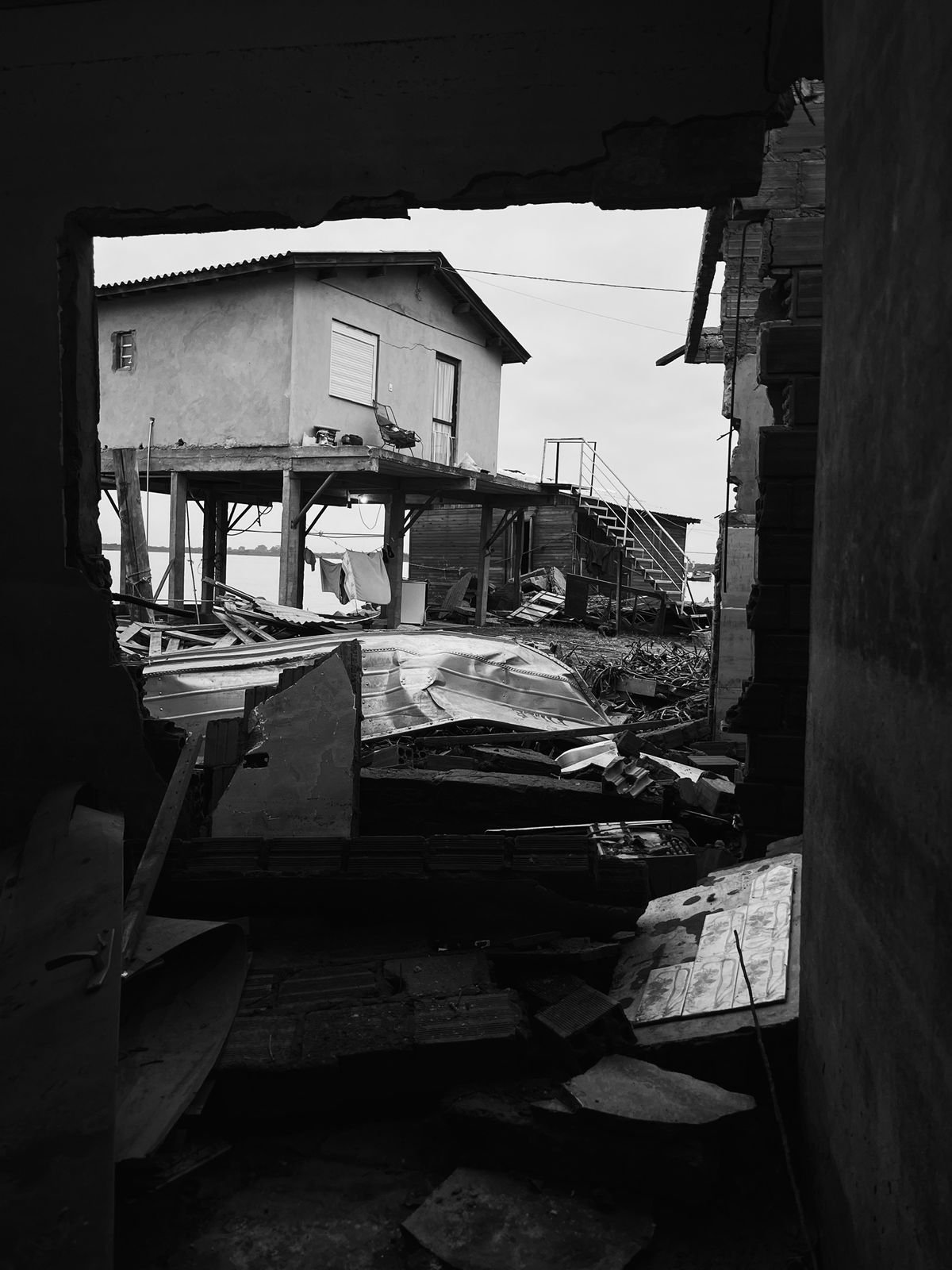 View of a damaged house in black and white, with debris, broken roof, and a house on stilts.