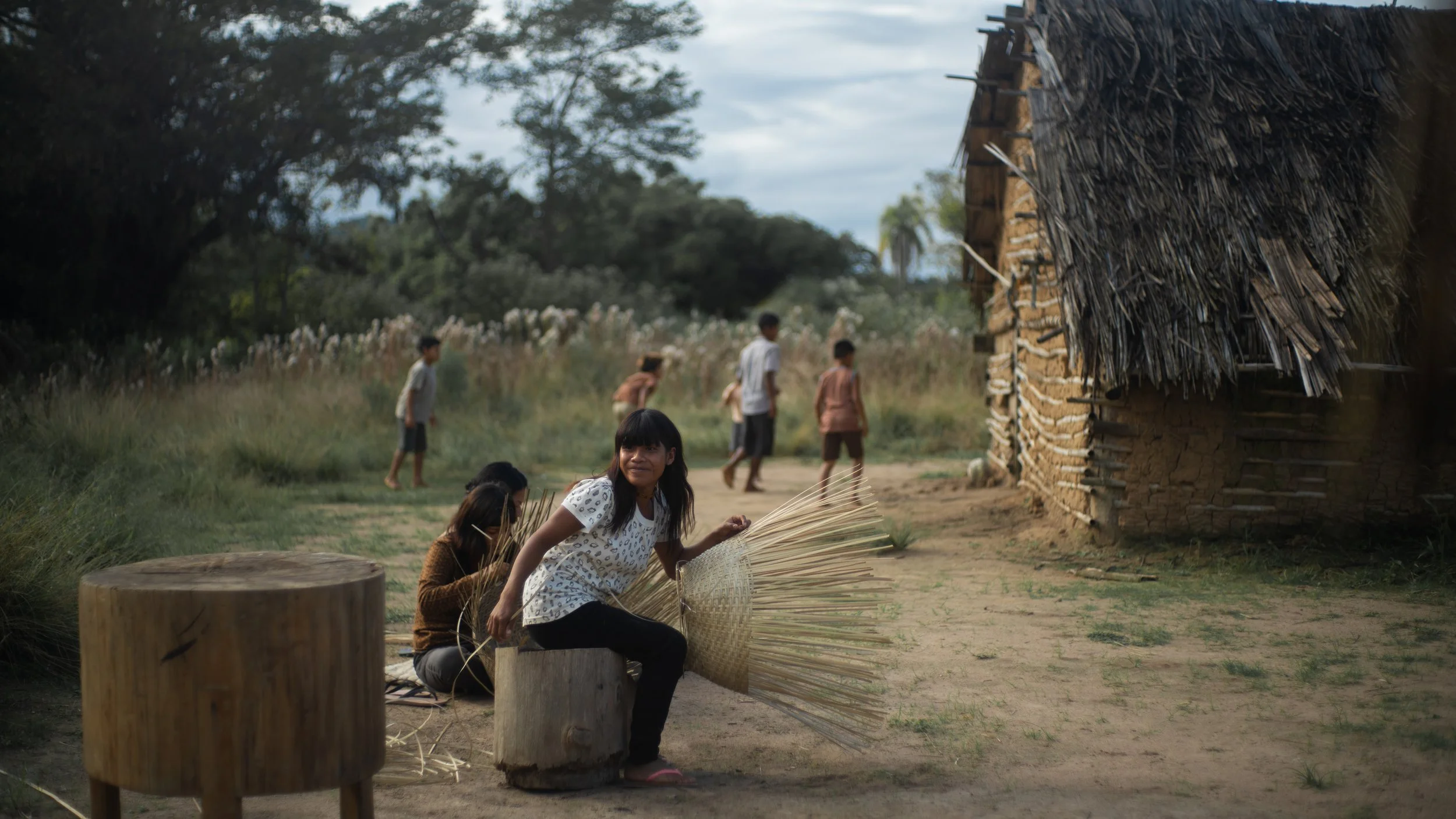 Children gather outside a rural house with a thatched roof, playing and weaving materials in a lush, green landscape.