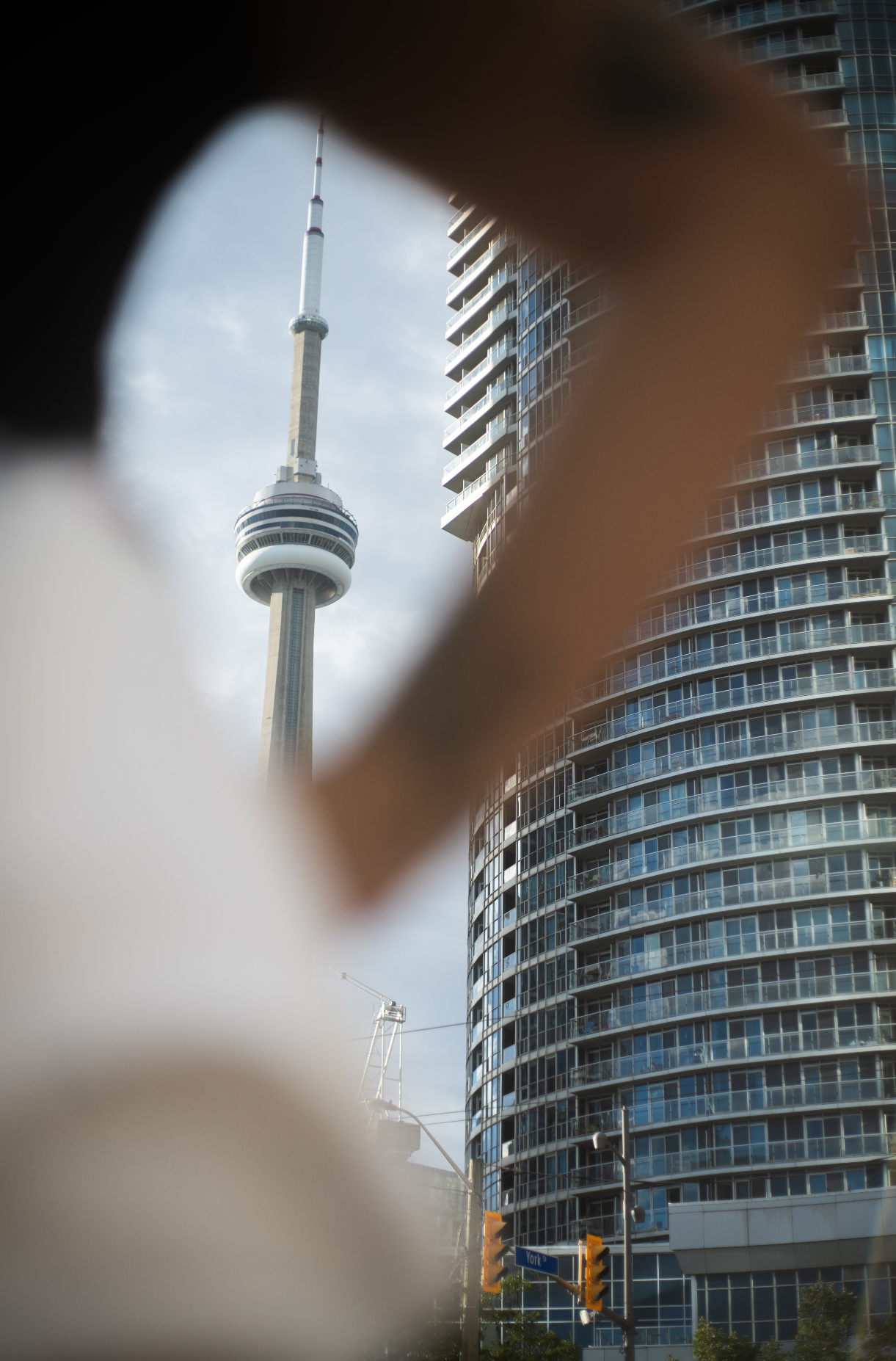 A cityscape with the CN Tower visible through a blurry, partially obstructed view, and a modern glass building with balconies on the right side. A street sign near the bottom reads 'York'.