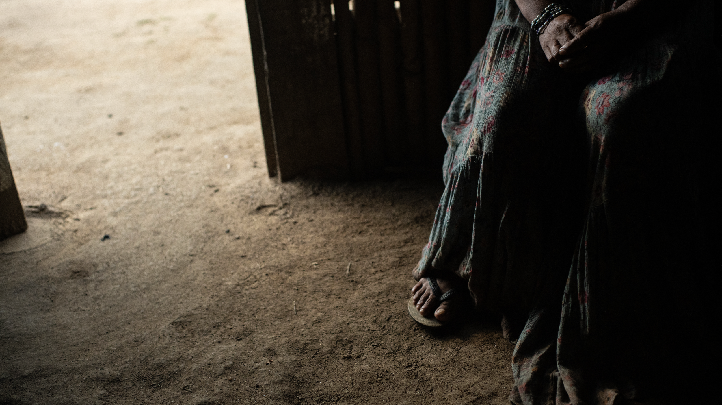Close-up of a person sitting with crossed legs, wearing patterned pants and flip-flops, in a dimly lit room with a dirt floor.