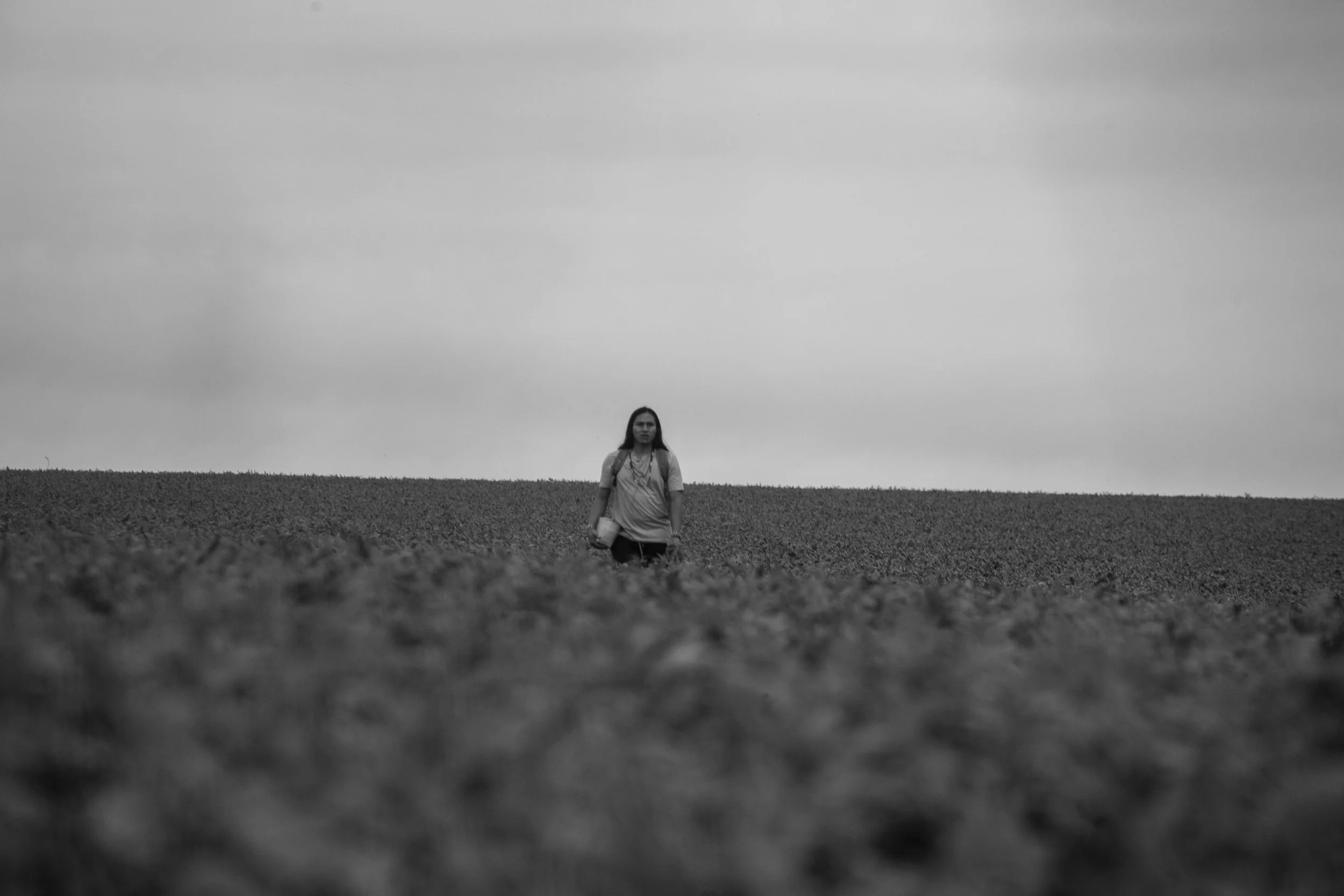 A black and white photo of a woman walking across a field with a cloudy sky in the background.