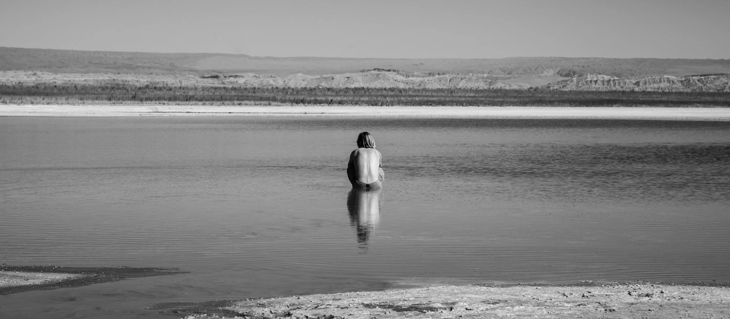 A person sitting on the edge of a body of water, with their back to the camera, surrounded by a landscape of mountains and open land in the distance.