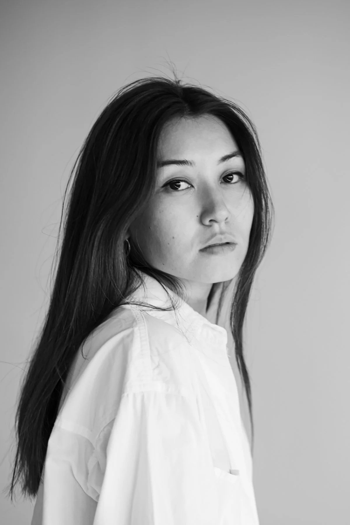 Black and white portrait of a woman with long hair, wearing a light-colored shirt, looking over her shoulder.