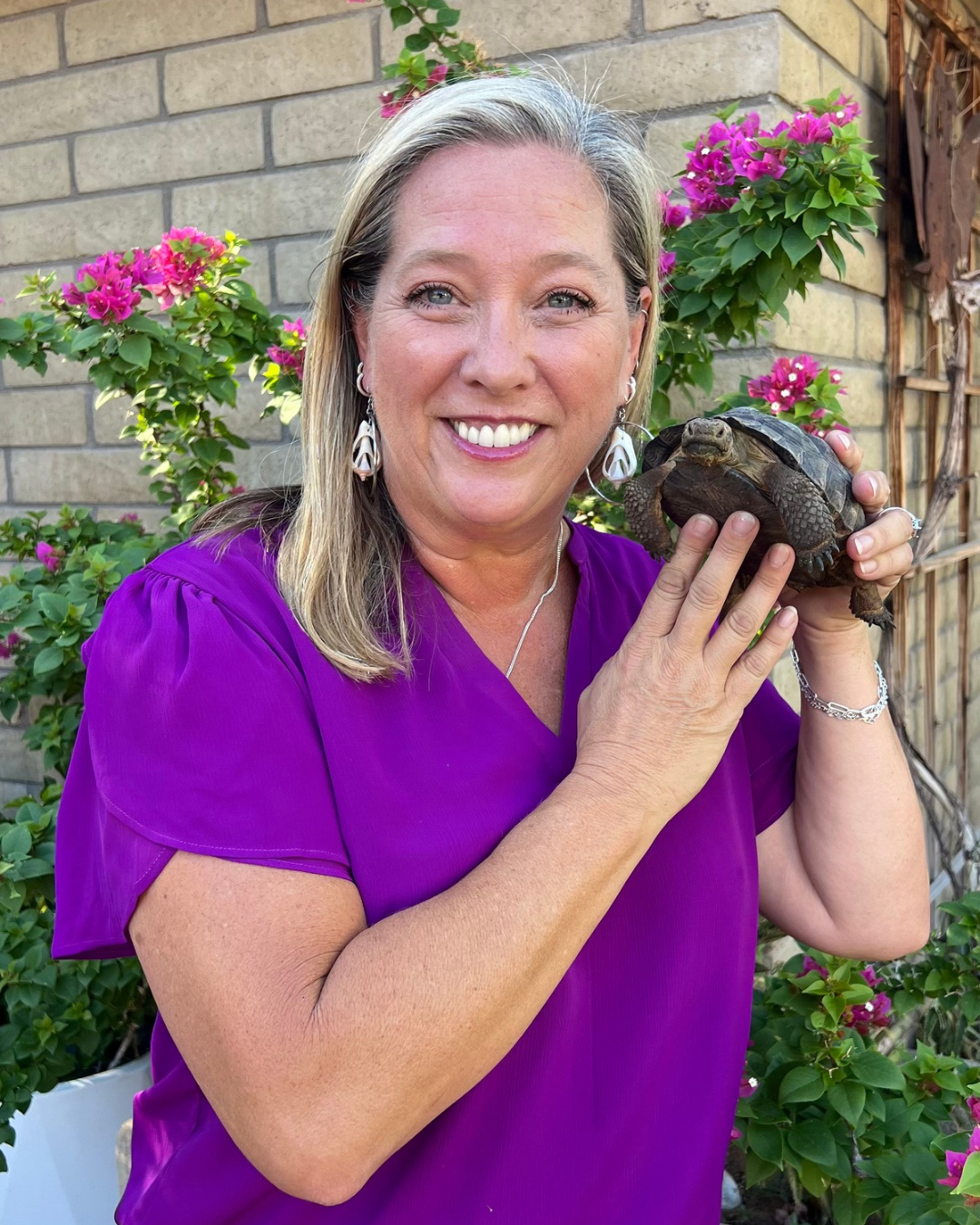 Local Laps owner Tara Lamar in a purple blouse holding a tortoise in front of flowering bougainvillea.