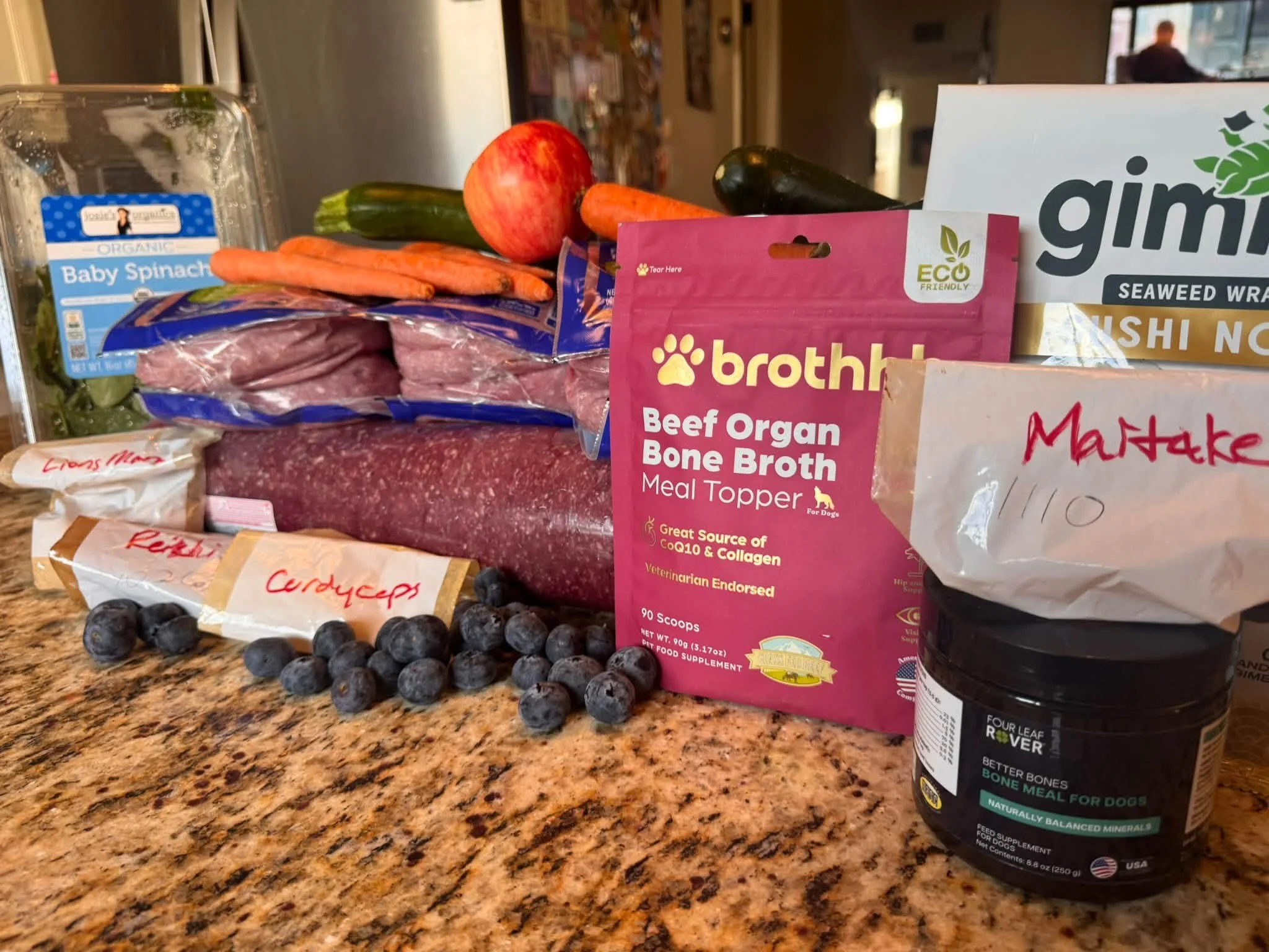 Groceries and dog supplements arranged on a kitchen counter, including spinach, carrots, apple, zucchini, eggplant, blueberries, ground meat, bone broth, and seaweed wraps.