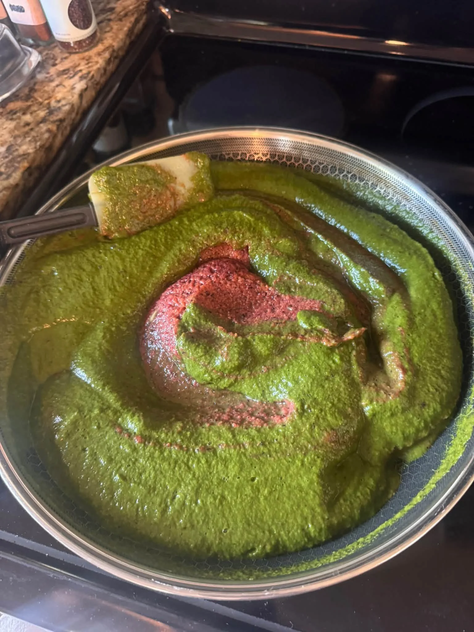 A pot of green curry sauce simmering on a stovetop with browned meat and a spatula.