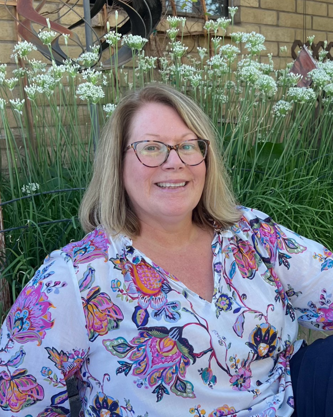 Lana from the Local Laps crew smiling in a colorful floral blouse and glasses, seating in the garden waiting for her next lap!